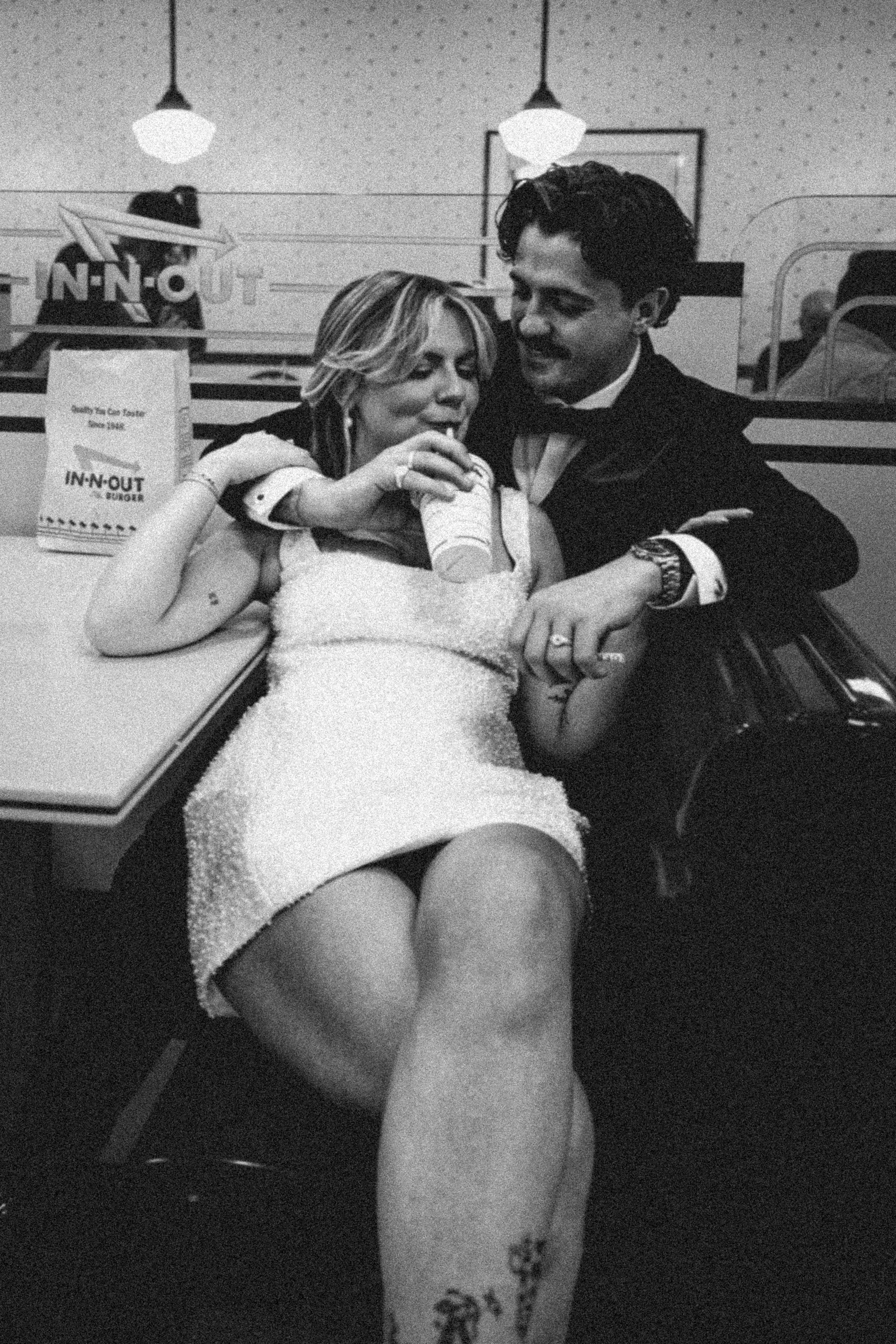 A black and white photo of a couple at an In-N-Out Burger restaurant, sitting at a table. The woman is wearing a strapless dress and is holding a cup with a straw, while the man is in a tuxedo. They are close together, smiling and looking at each oth