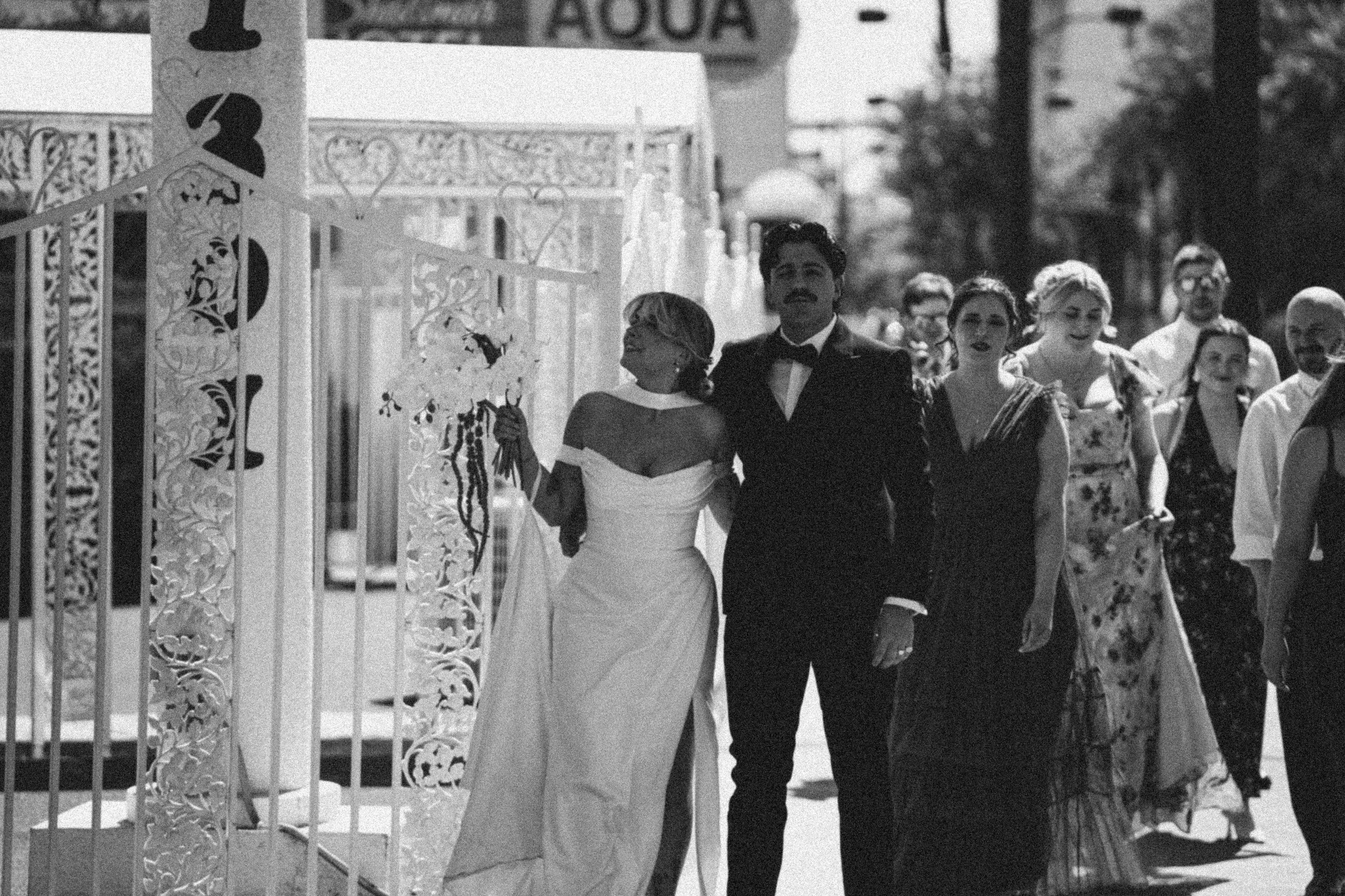Black and white photo of a bride and groom walking through a gate, surrounded by a group of people at a wedding.