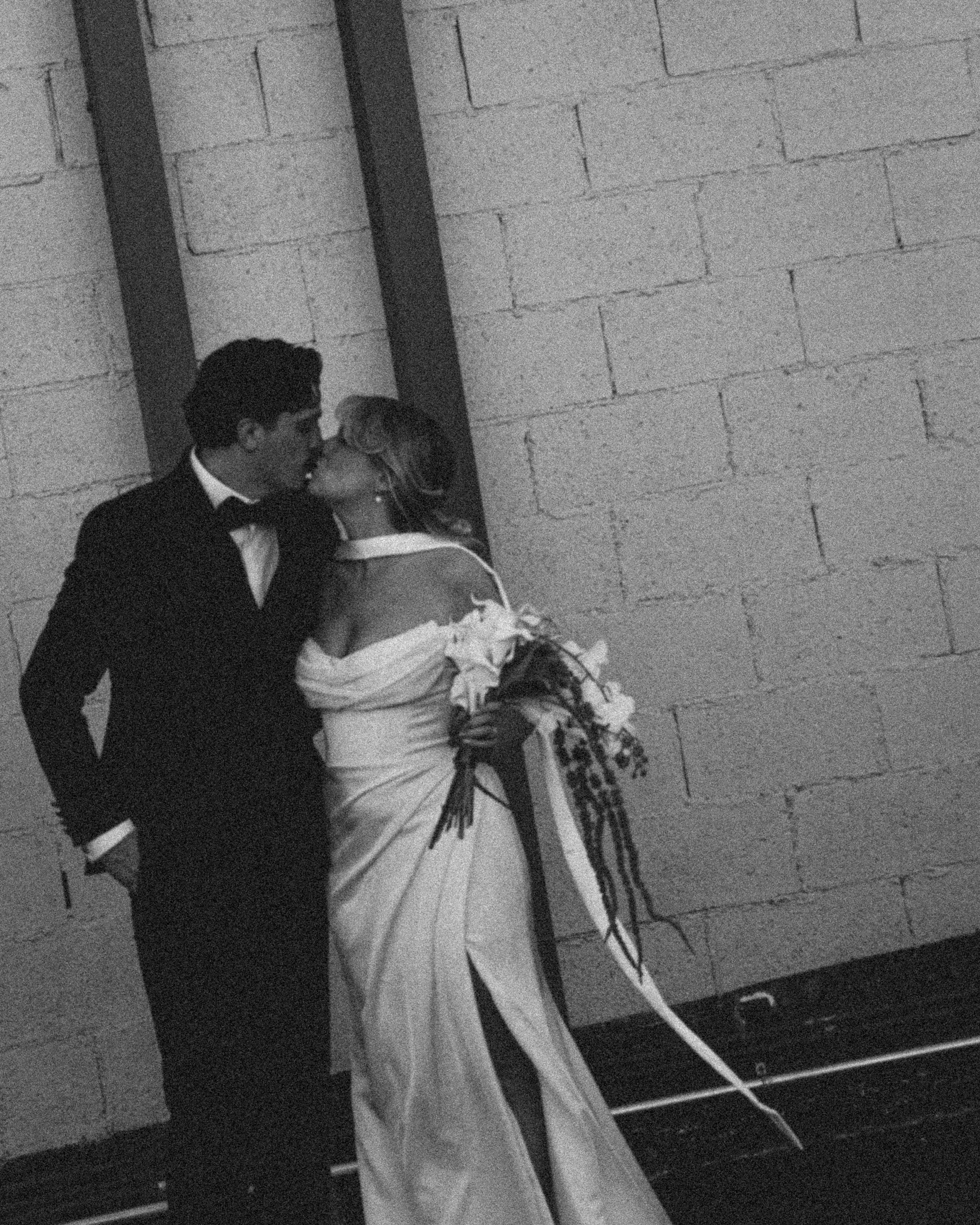 A couple dressed in formal attire, with the man in a tuxedo and the woman in an elegant dress, sharing a kiss in front of a decorated table with white cloth, flowers, and ribbons, likely at a celebration or event.