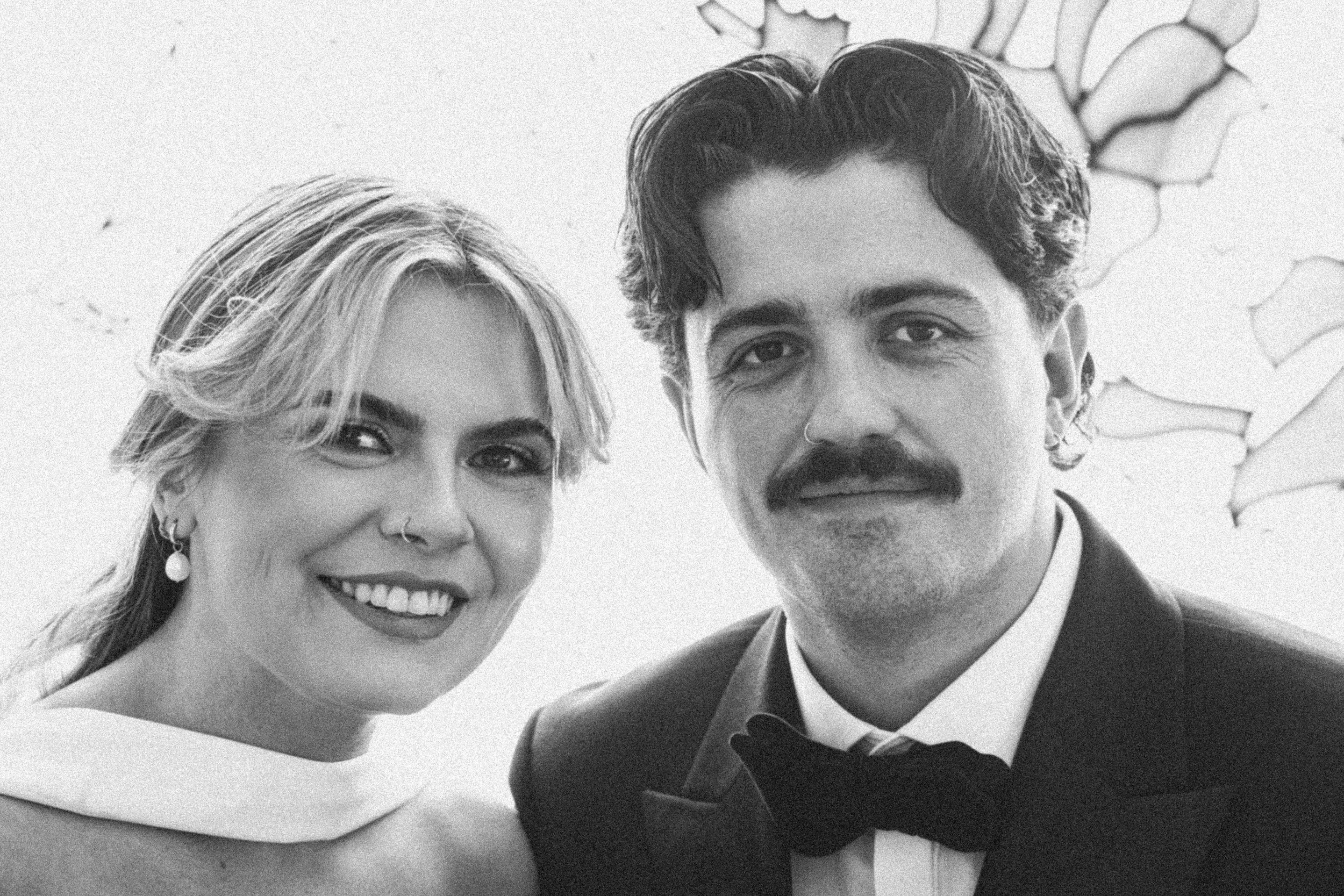 Black and white photo of a smiling woman with blonde hair and earrings, and a man with curly hair and a bow tie, standing close together.