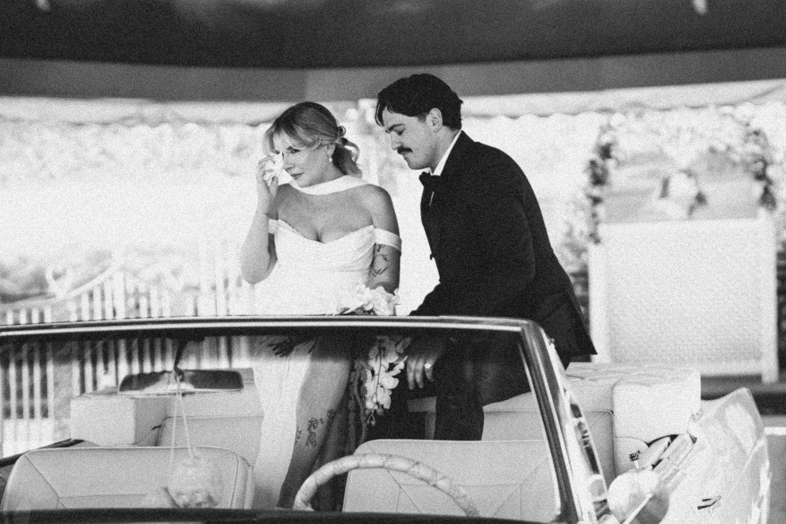 A black-and-white photo of a bride and groom in wedding attire standing next to a car. The bride has her hand near her face, holding a tissue or handkerchief, and the groom is looking down. The scene appears to be outdoors with blurred trees in the b