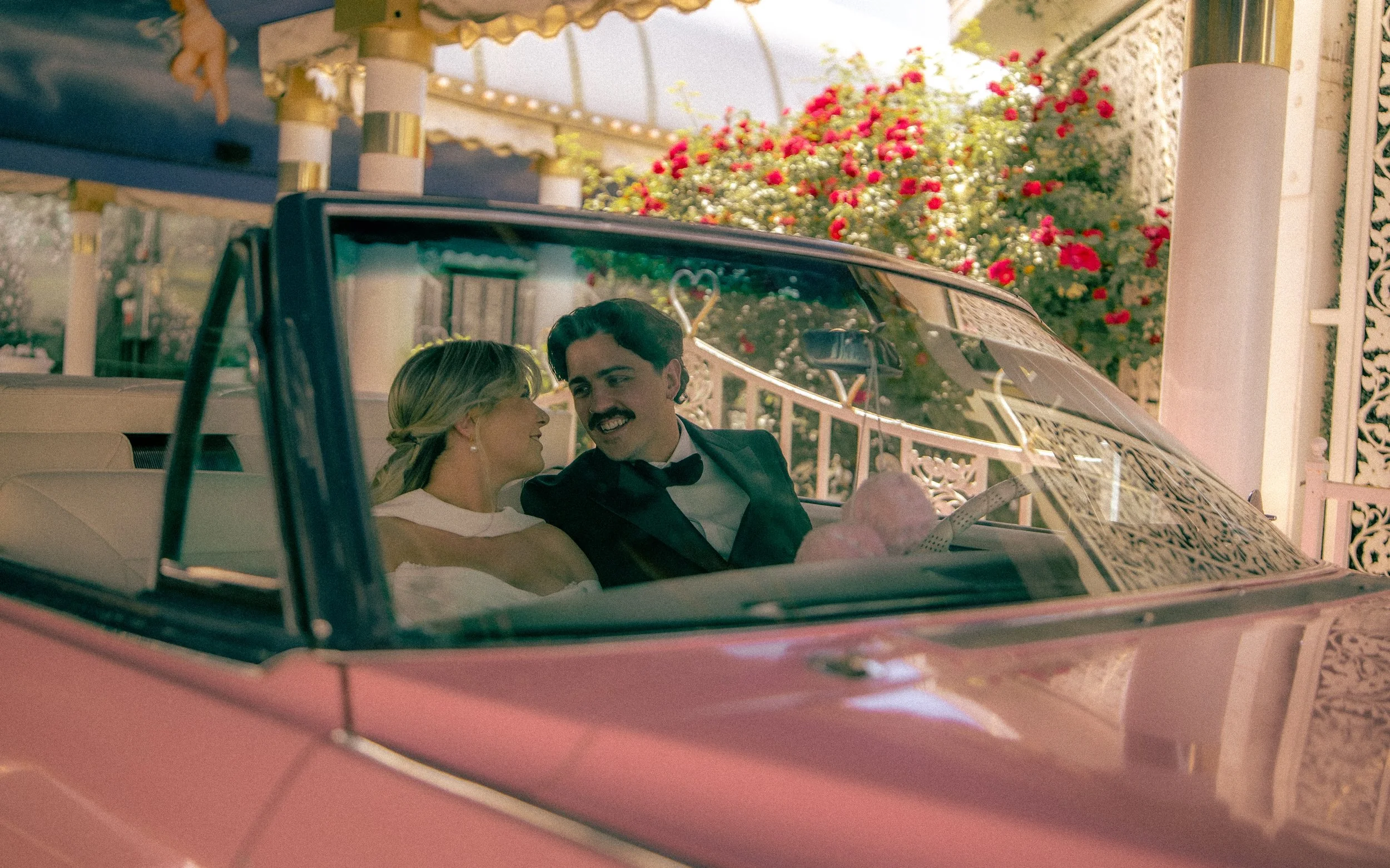 A couple dressed in formal wedding attire, sitting in a pink vintage car, smiling and looking at each other. The background features lush greenery and pink flowers, suggesting a wedding or special event setting.