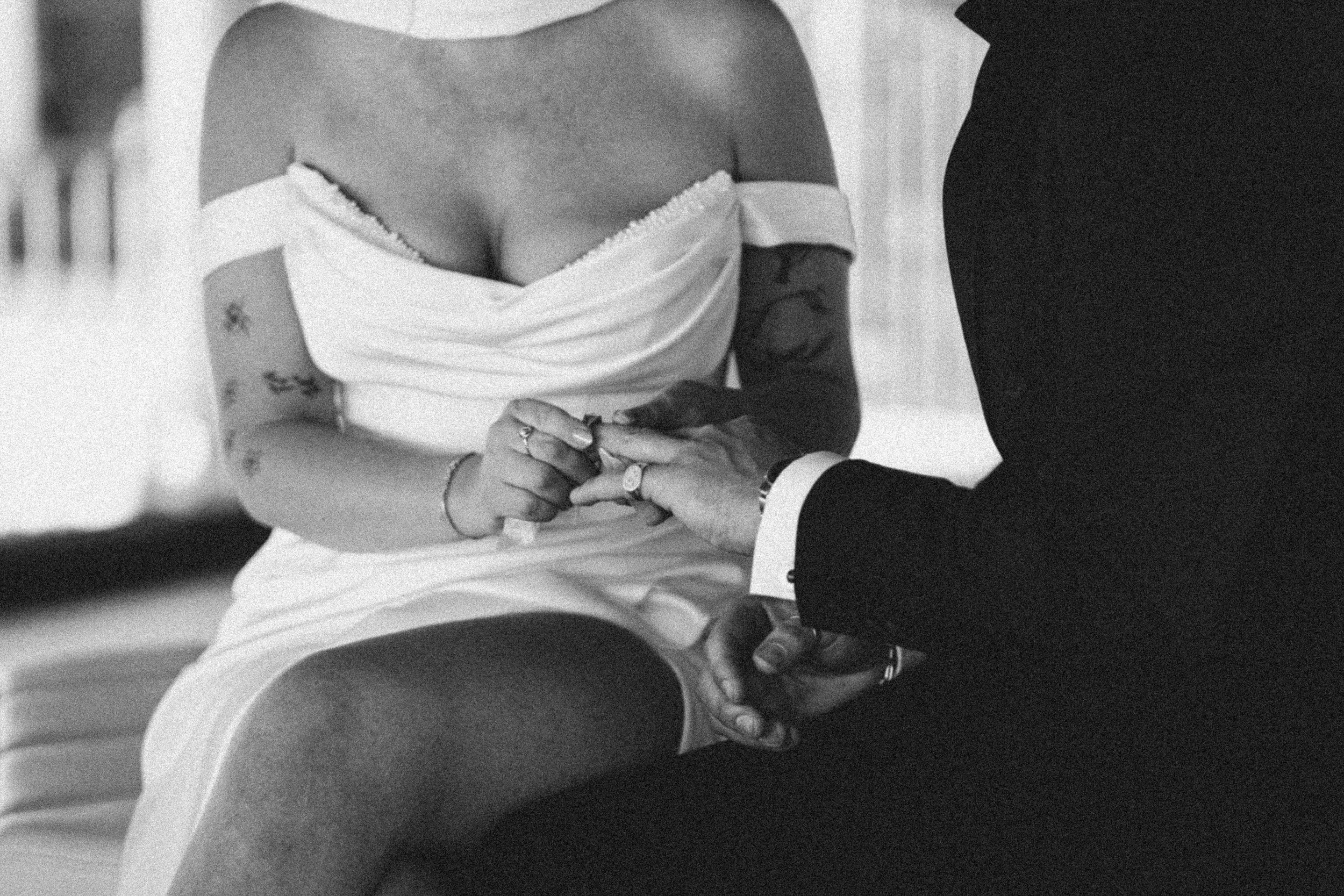 A black-and-white photo of a person in formal attire, holding hands with another person. The person appears to be wearing a white off-the-shoulder dress with a tattoo on their arm and a ring on their finger. The other person is dressed in a dark suit.