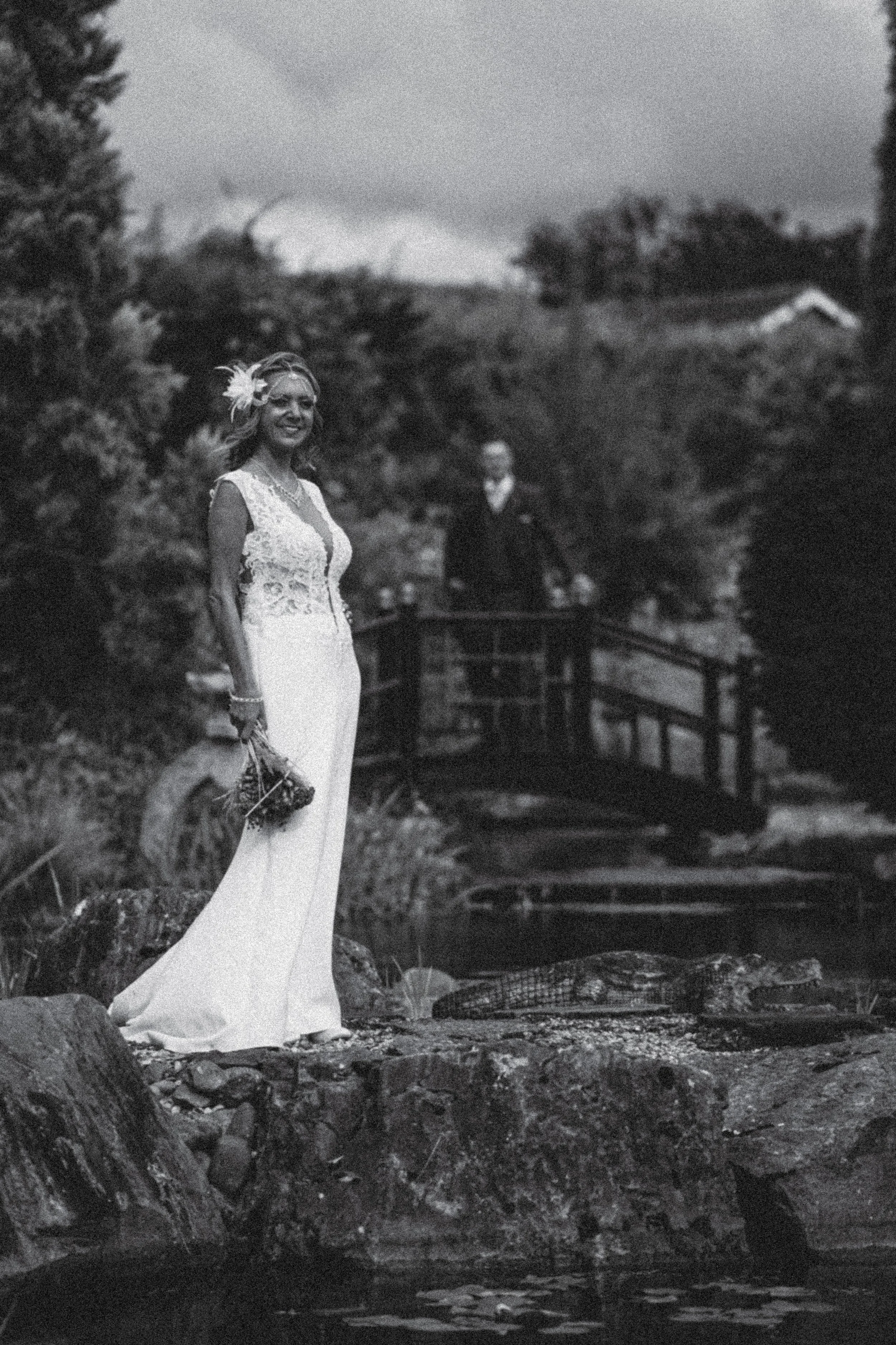 A woman in a wedding dress stands near a stream with a smiling expression, a man is in the background on a bridge, and a landscape of trees, a house, and mountains is visible in the evening or nighttime setting.