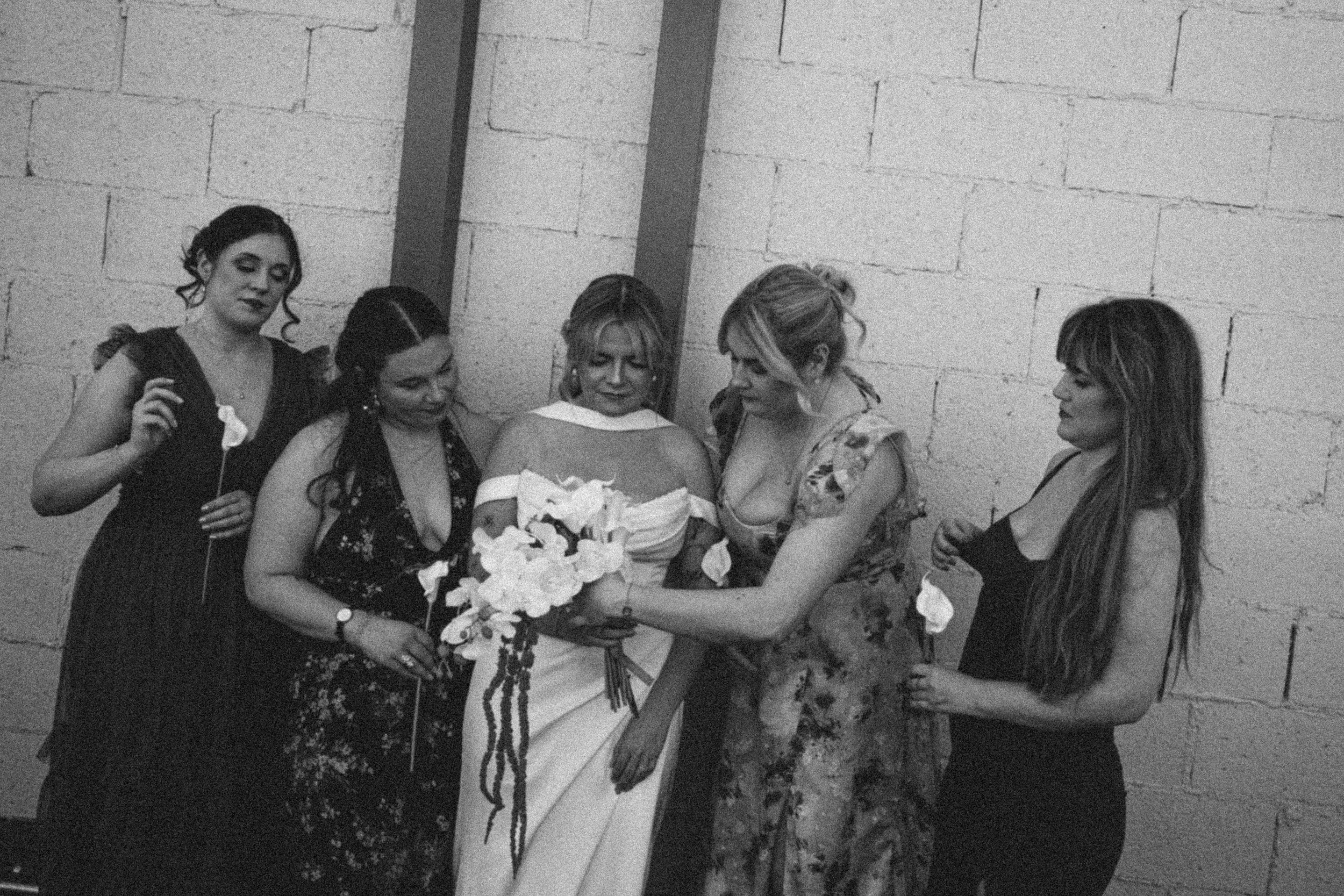 Group of five women standing against a brick wall, with one woman in a white dress holding a bouquet of flowers, possibly celebrating a special occasion.