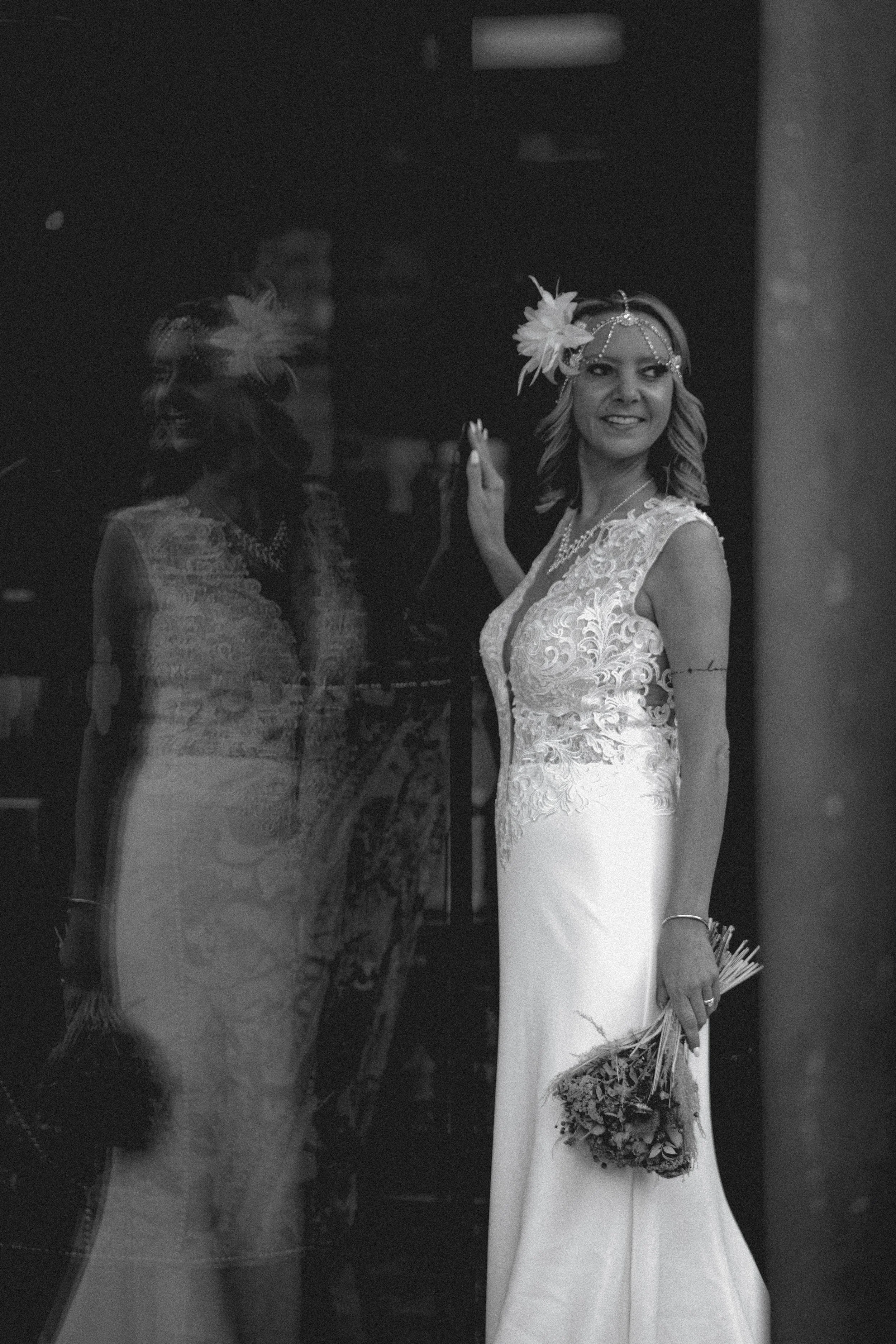 Bride in a lace wedding dress with a floral headpiece and holding a bouquet, giving a smile while looking through a glass reflection.