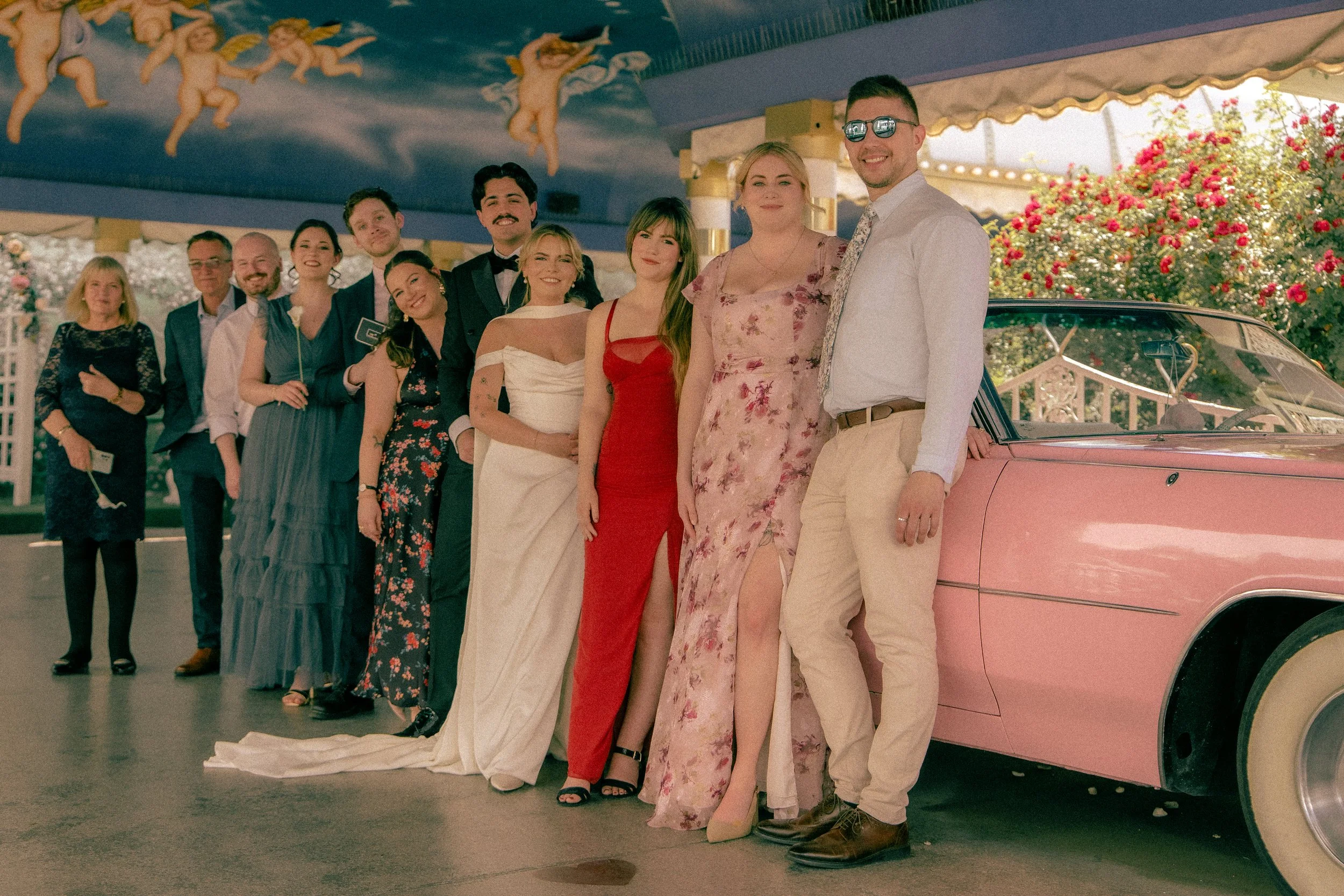 Group of people posing for a photo in front of a pink vintage car during a celebration or wedding event, with a decorated ceiling featuring cherubs and a background of flowering bushes.