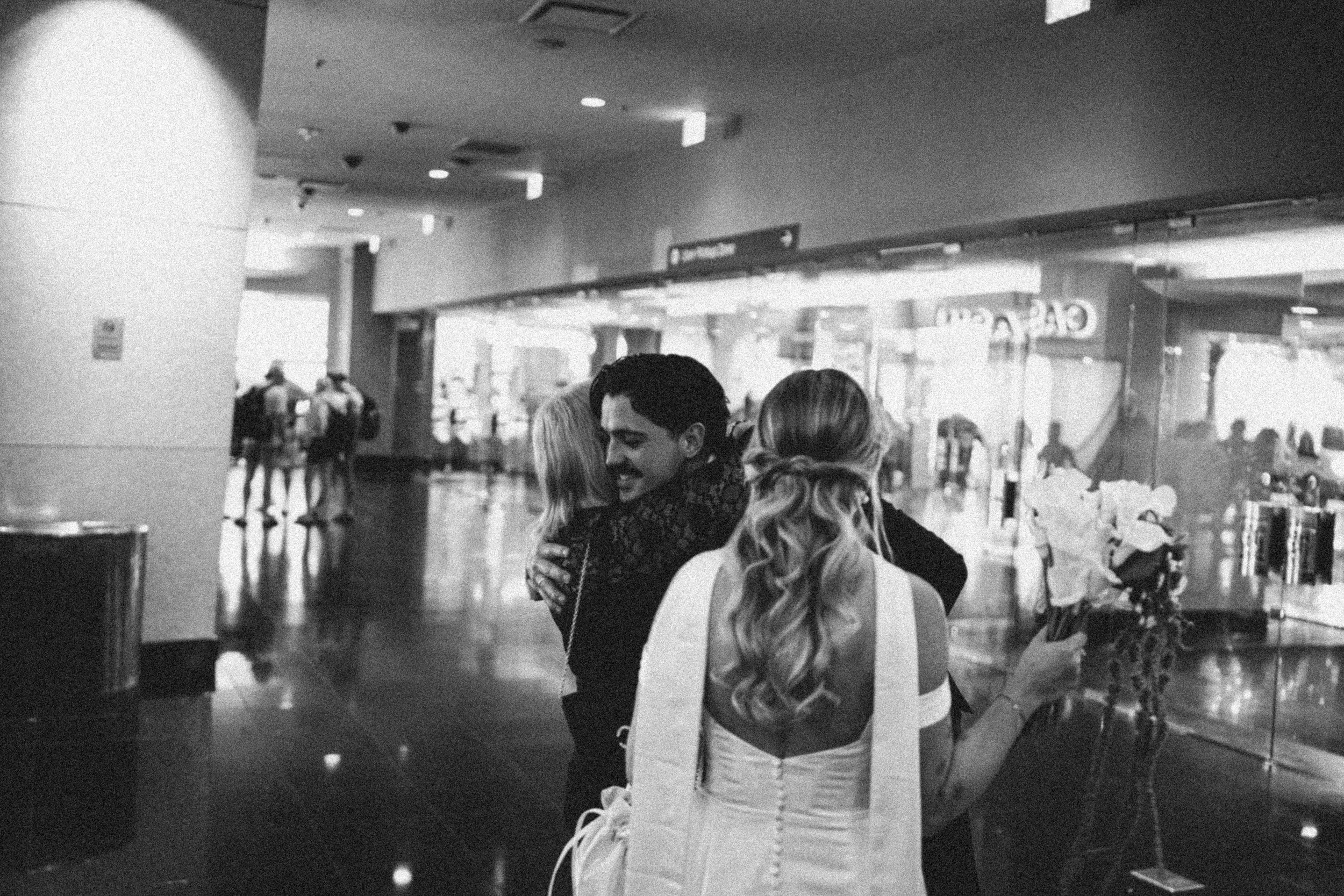 People in an airport terminal, with a man hugging a woman in the foreground and other travelers waiting or walking in the background.