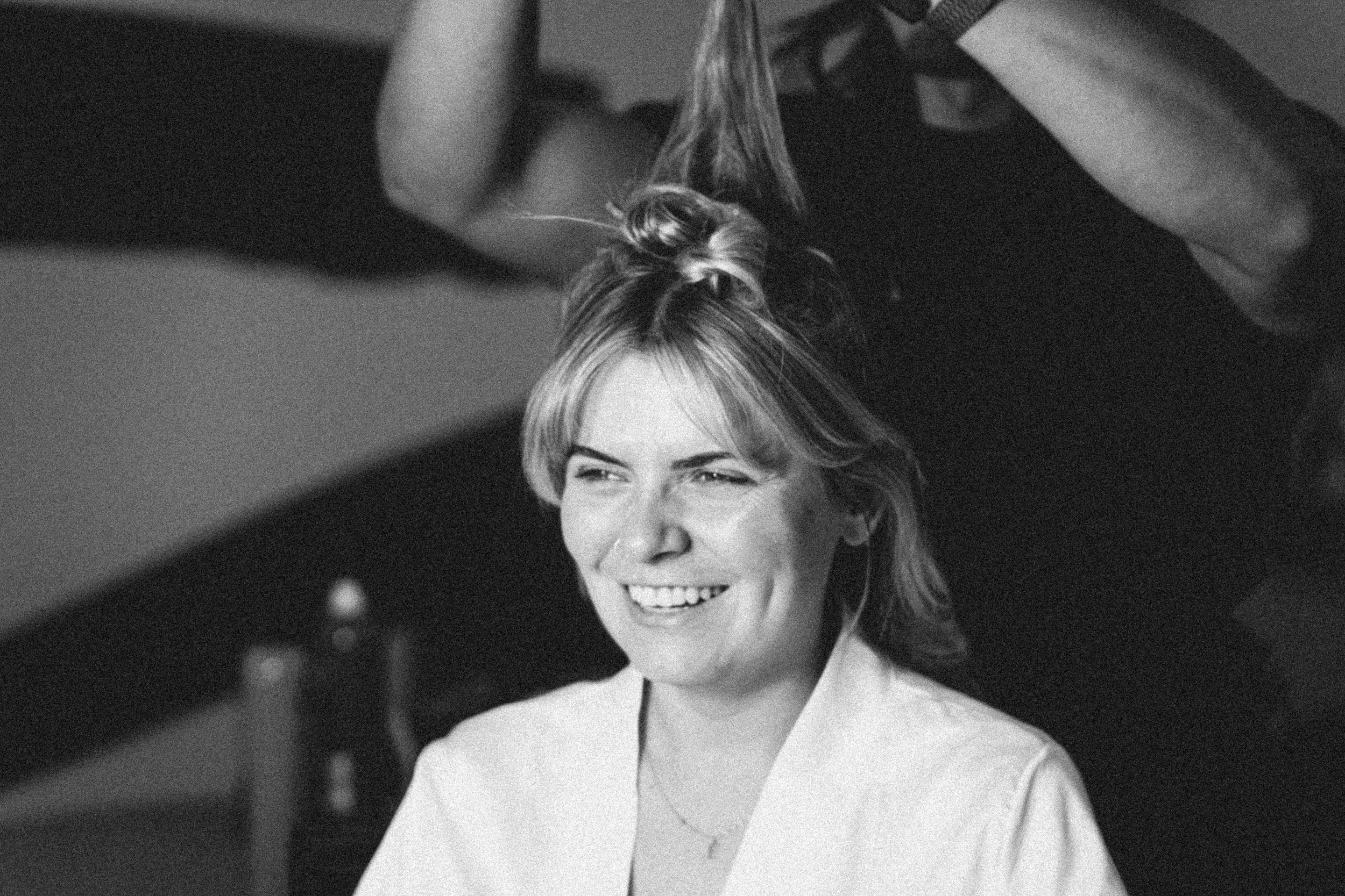 Black and white photo of a woman with short, wavy hair smiling as a person styles her hair, out of focus in the foreground.