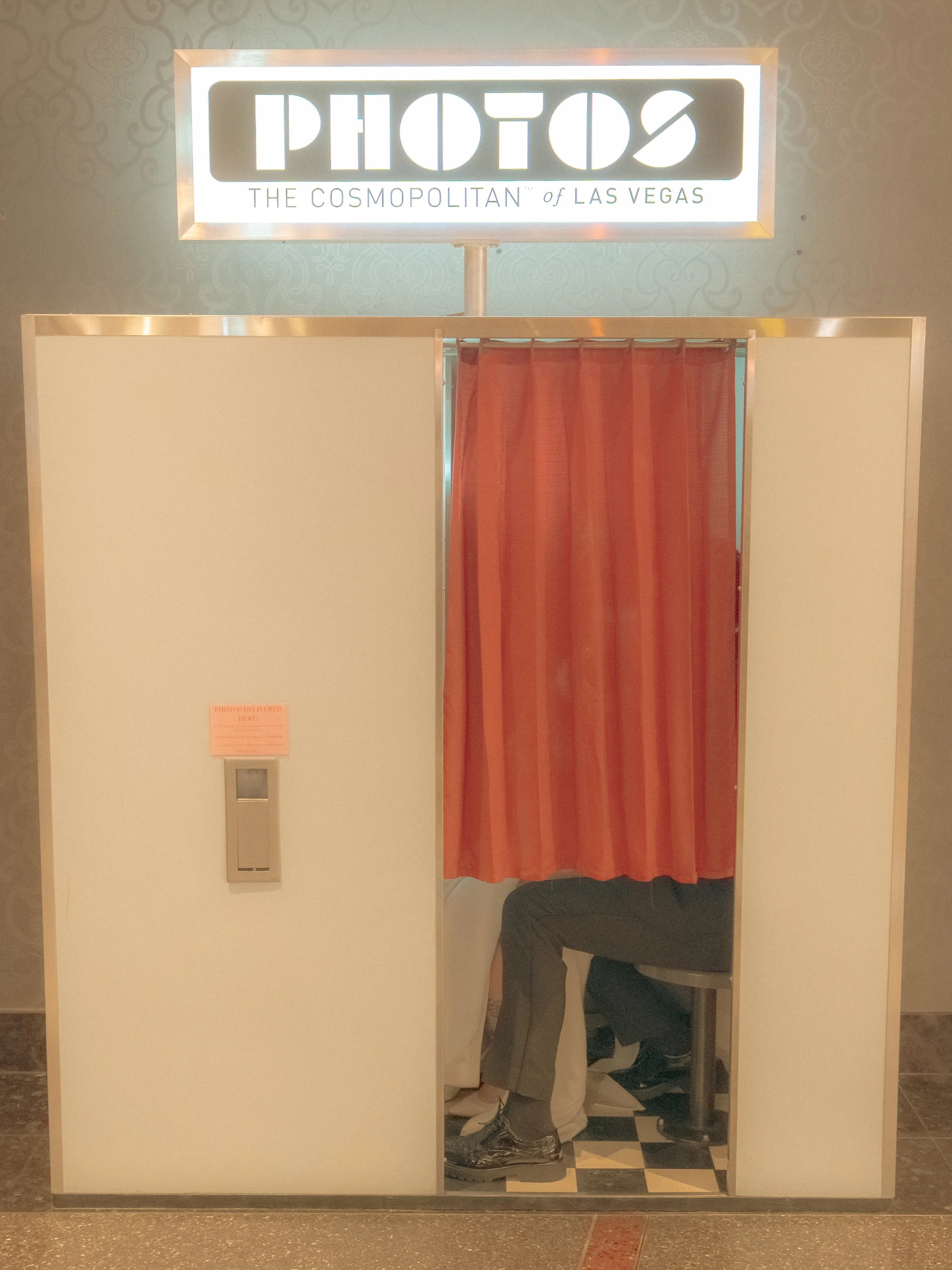 A person taking a photo of themselves in a vanity mirror booth at a photo booth station labeled 'PHOTOS - THE COSMOPOLITAN of LAS VEGAS', with a red curtain inside the booth and a small sign on the left side of the booth.