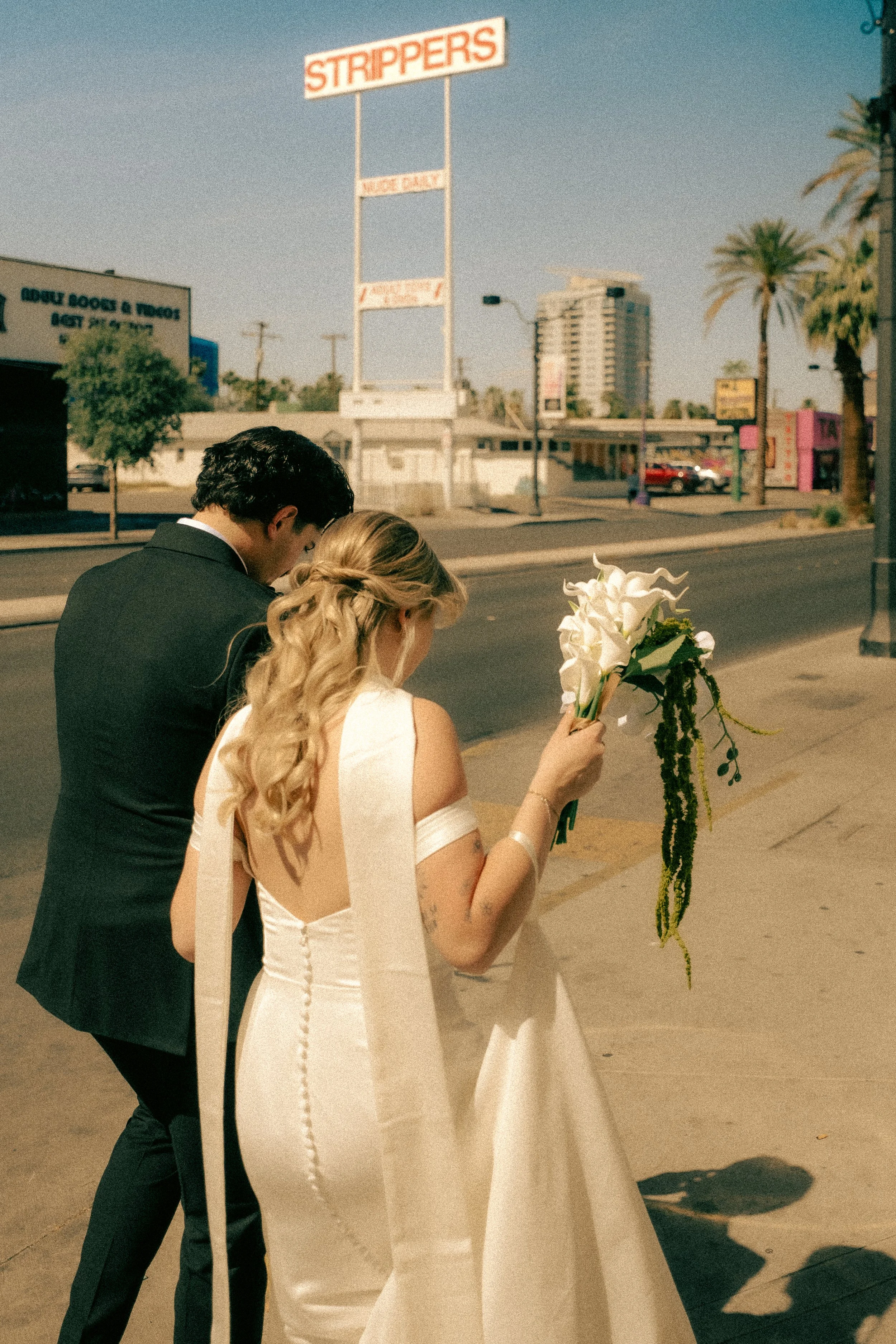 A bride and groom are standing next to each other outside, near a white display board. The bride is wearing a white wedding dress with buttons down the back and holding a bouquet of white flowers and greenery. The groom is dressed in a black suit. Th