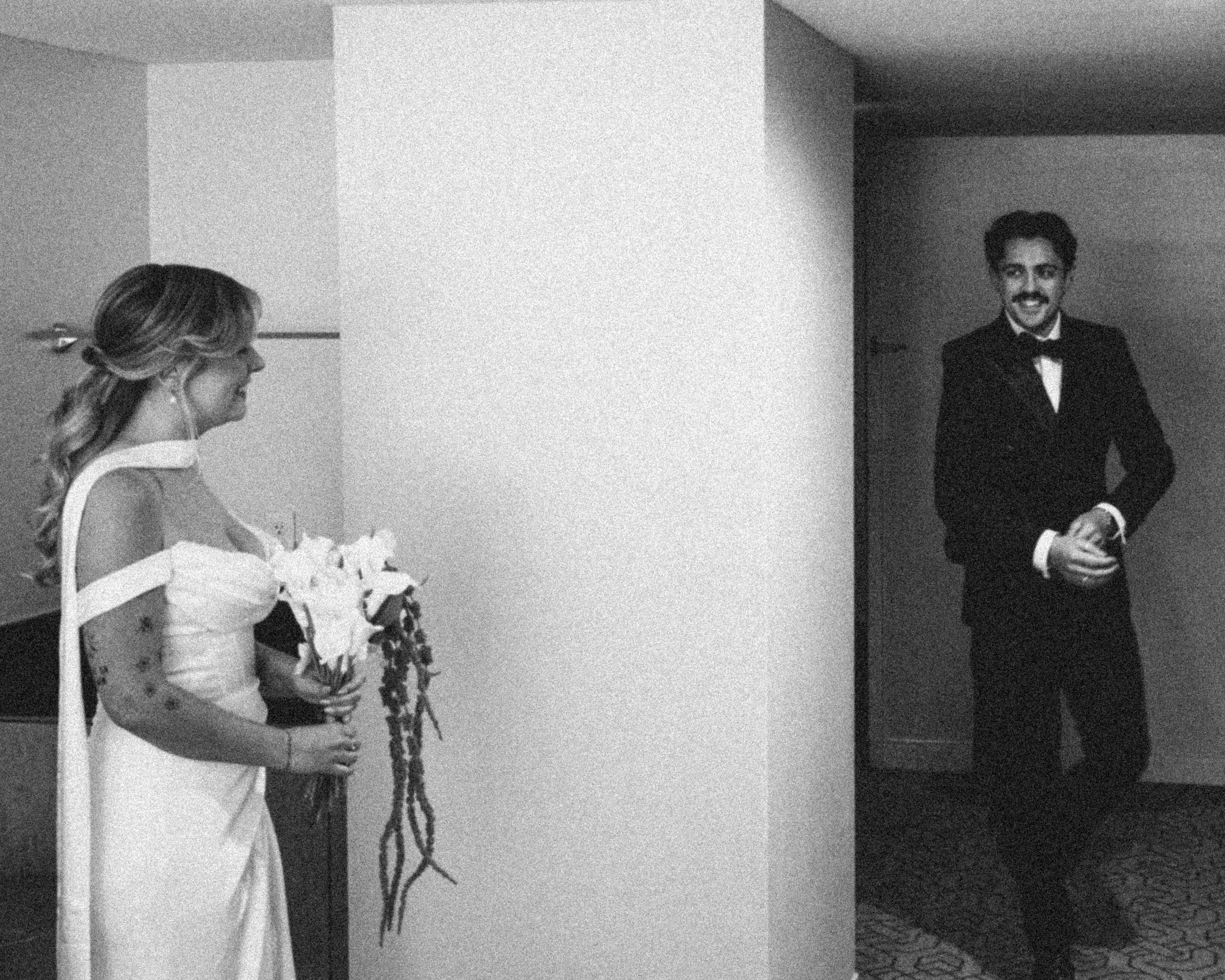 A woman in a white dress holding a bouquet of flowers and a man in a tuxedo with a boutonnière, standing in a room with plain walls, possibly at a wedding or formal event.