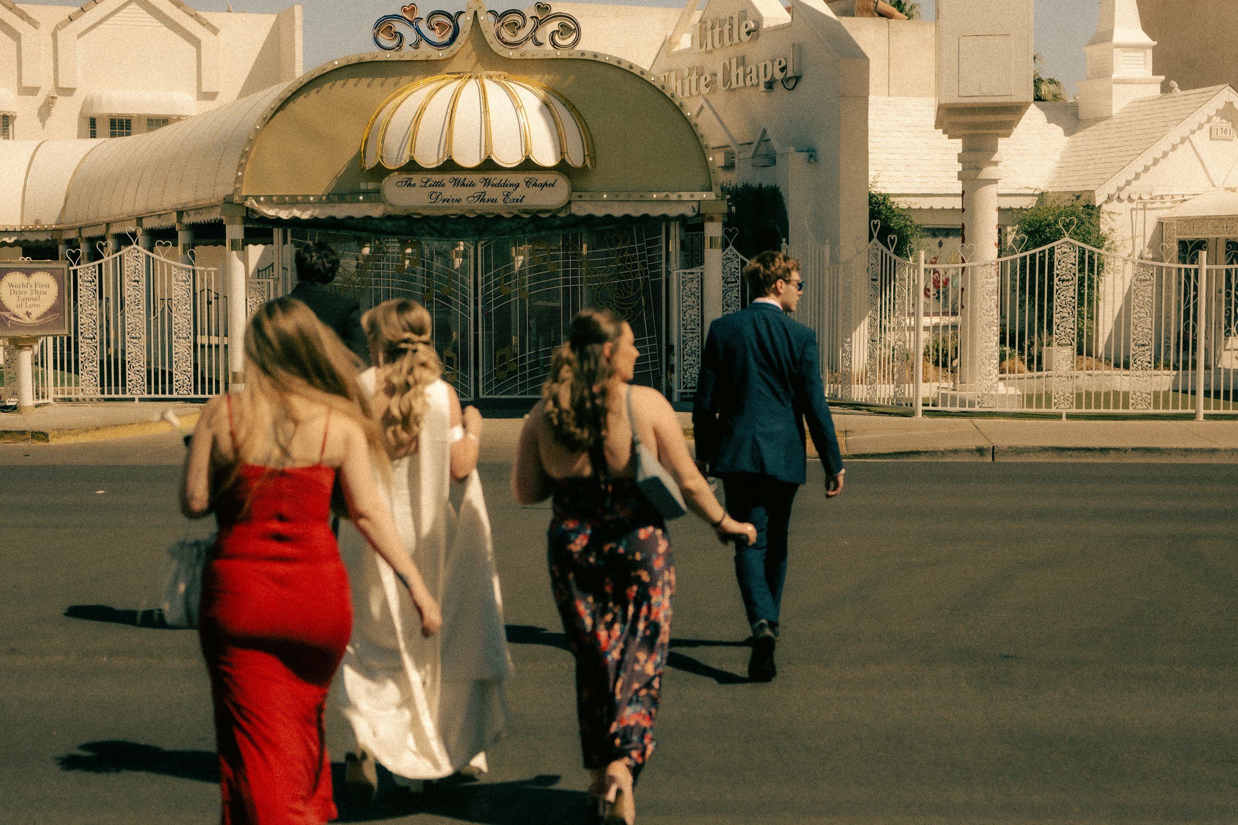 People walking on the street near the Little White Wedding Chapel, which has a drive-thru exit and decorative fencing, in daylight.