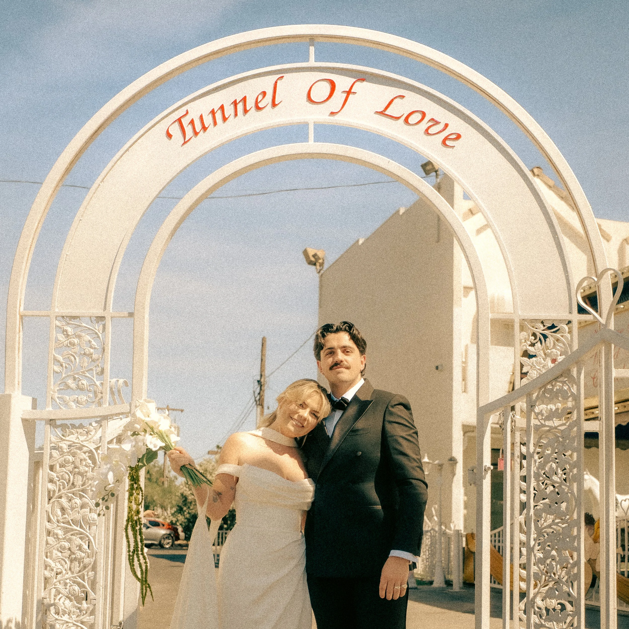 A couple dressed in wedding attire standing under a decorative white arch with the words "Tunnel of Love" written in red above them. The bride is holding a bouquet of white flowers, and they are smiling.