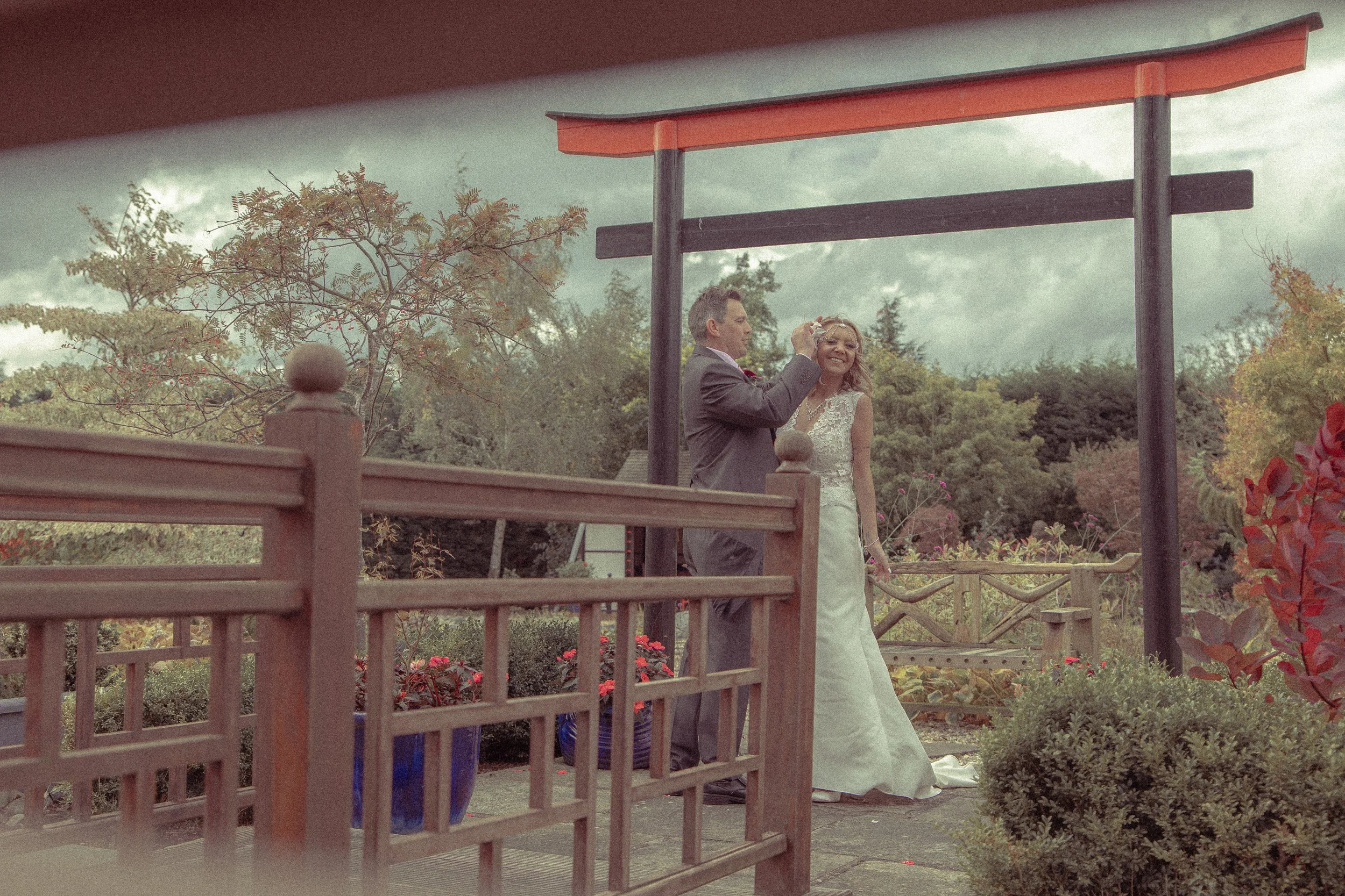 A bride and groom standing outdoors near a traditional Japanese torii gate, with trees and colorful plants in the background, under a cloudy sky.