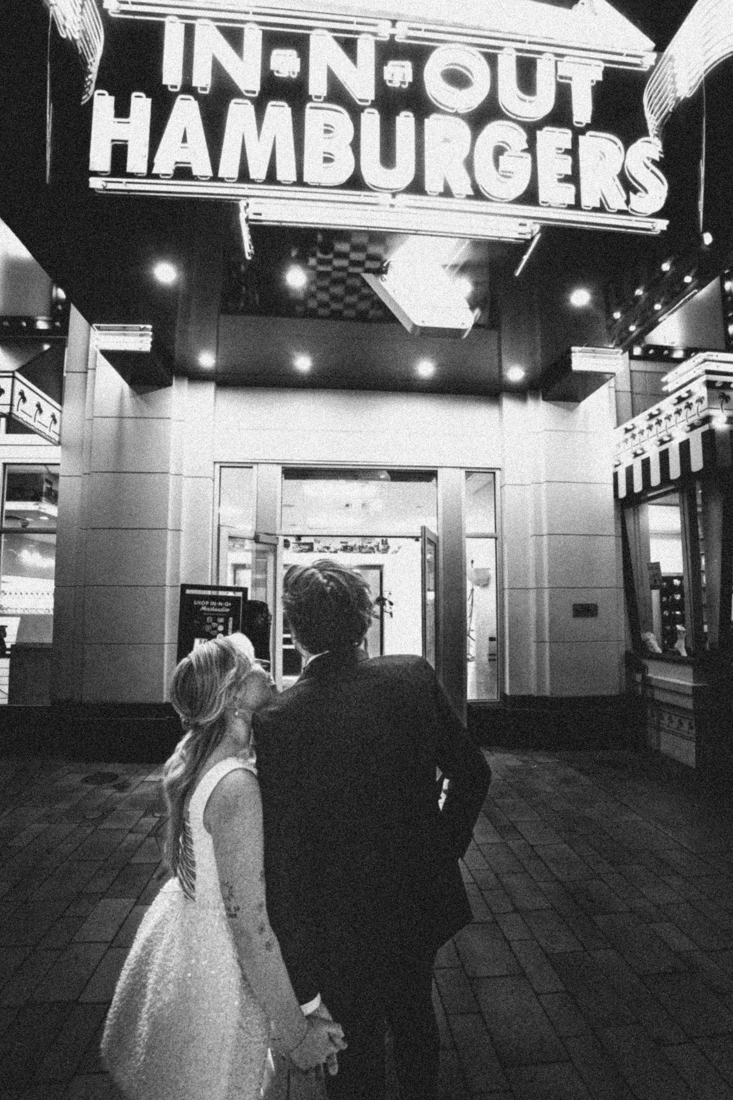 A man and a woman stand close together in front of a restaurant named "Inn Out Hamburgers" at night. The woman has long blonde hair, and the man has short hair. They appear to be looking at each other or at something nearby.