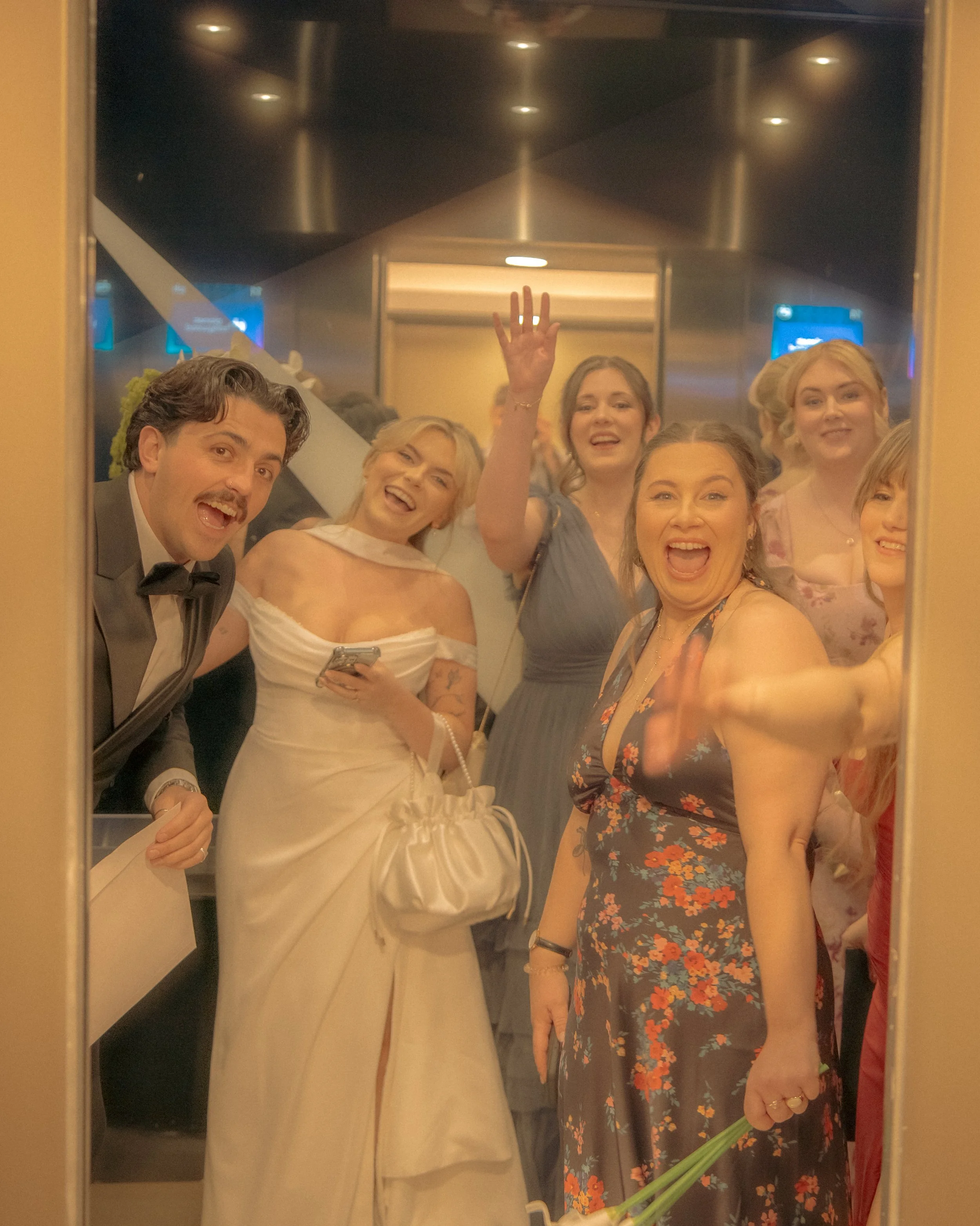 People taking a selfie in an elevator mirror at a social event, with one woman holding flowers and others smiling and waving.