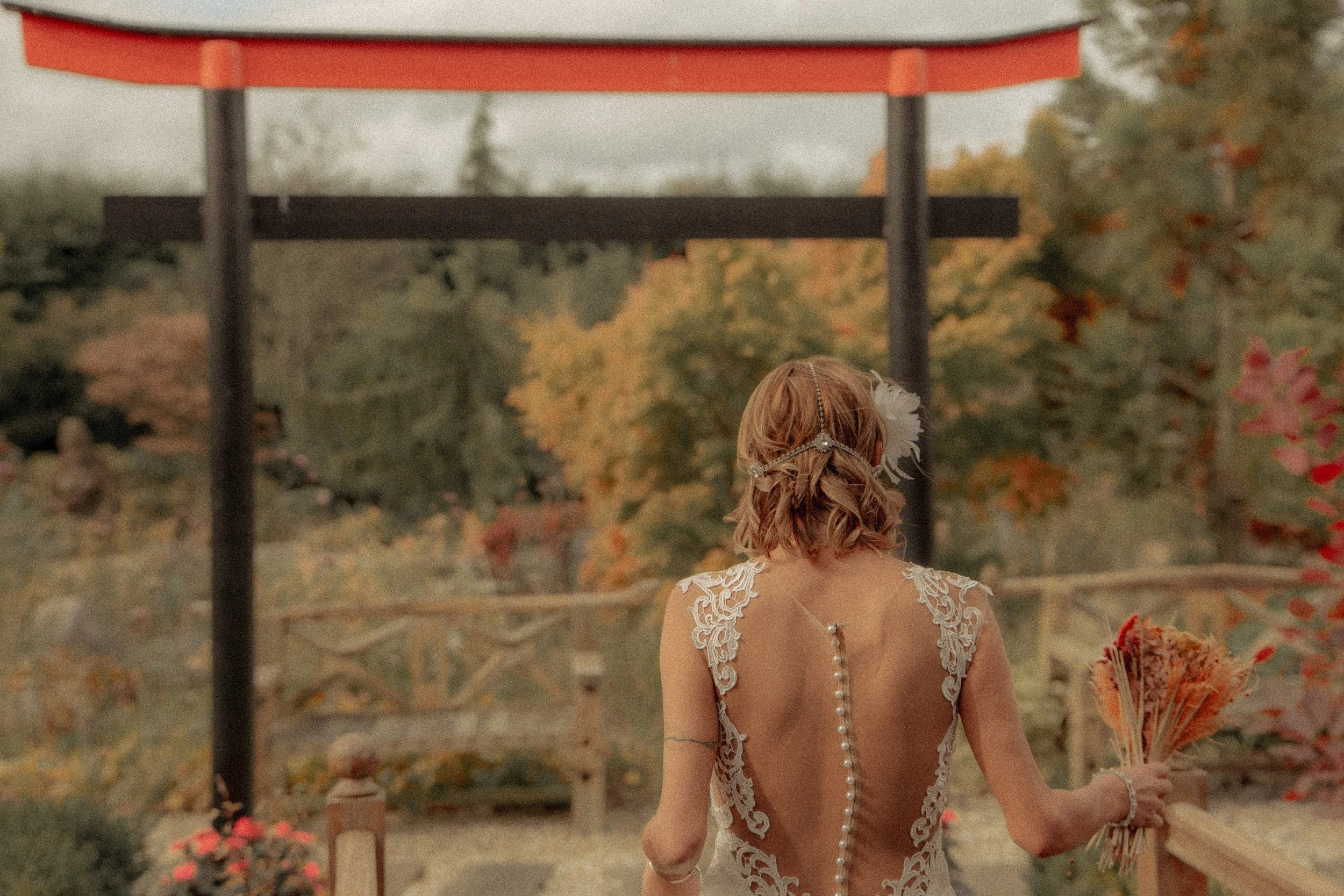 A woman with light brown, curled hair dressed in a backless lace wedding gown with pearl details, holding a bouquet, at an outdoor wedding venue with trees and a wooden altar in the background.
