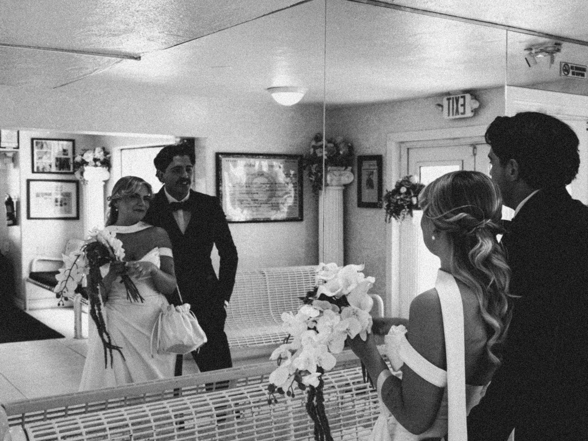A wedding ceremony taking place indoors with a bride, groom, and guests. The bride is wearing a wedding dress holding a bouquet, and the groom is standing beside her with a woman, possibly a family member or officiant. The room is decorated with flow