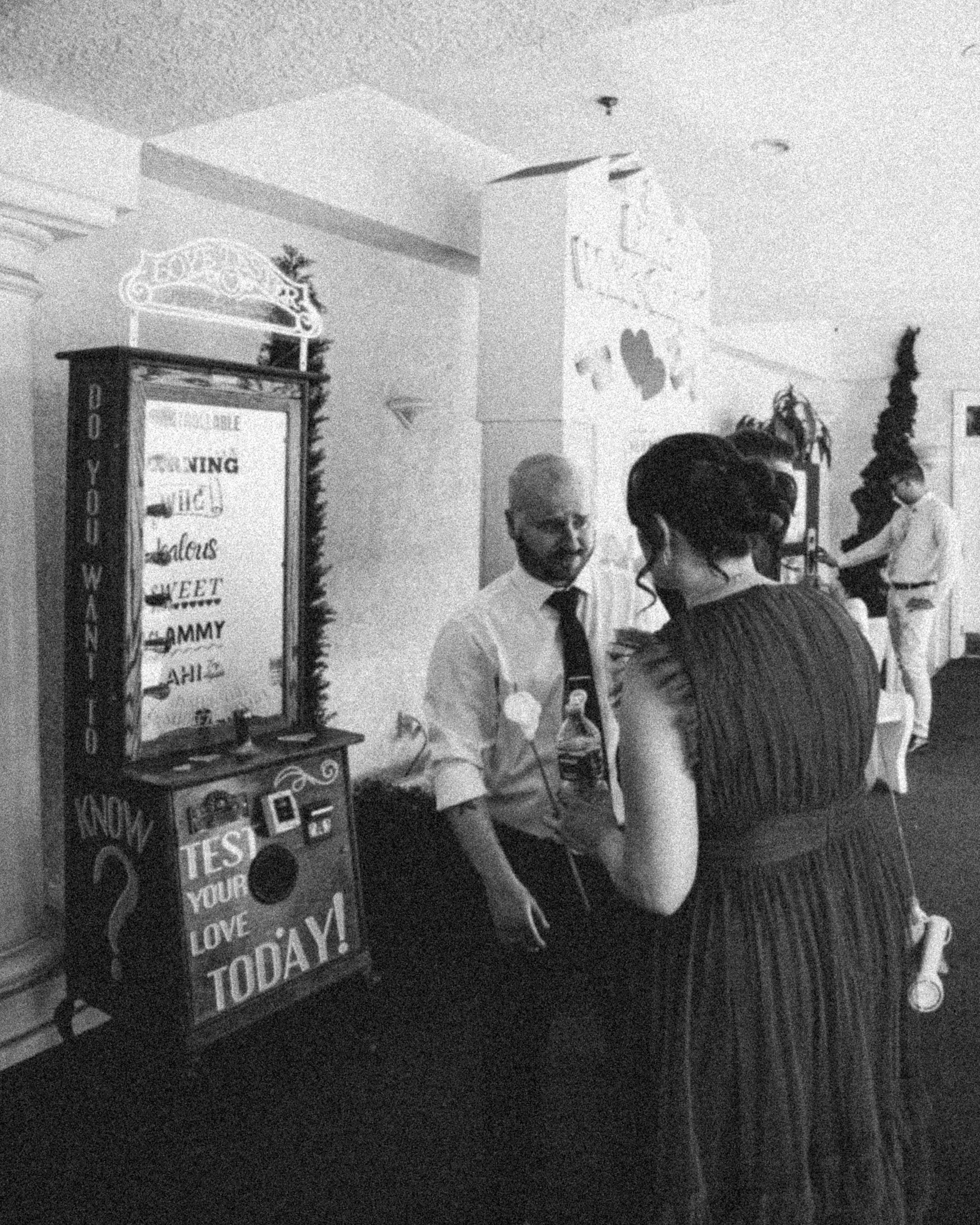 A group of people in a room with a large sign that reads 'We are open' and a menu listing food items including waffles, pancakes, and coffee. One person with glasses and a tie is talking to a woman with her back to the camera. There are holiday decor