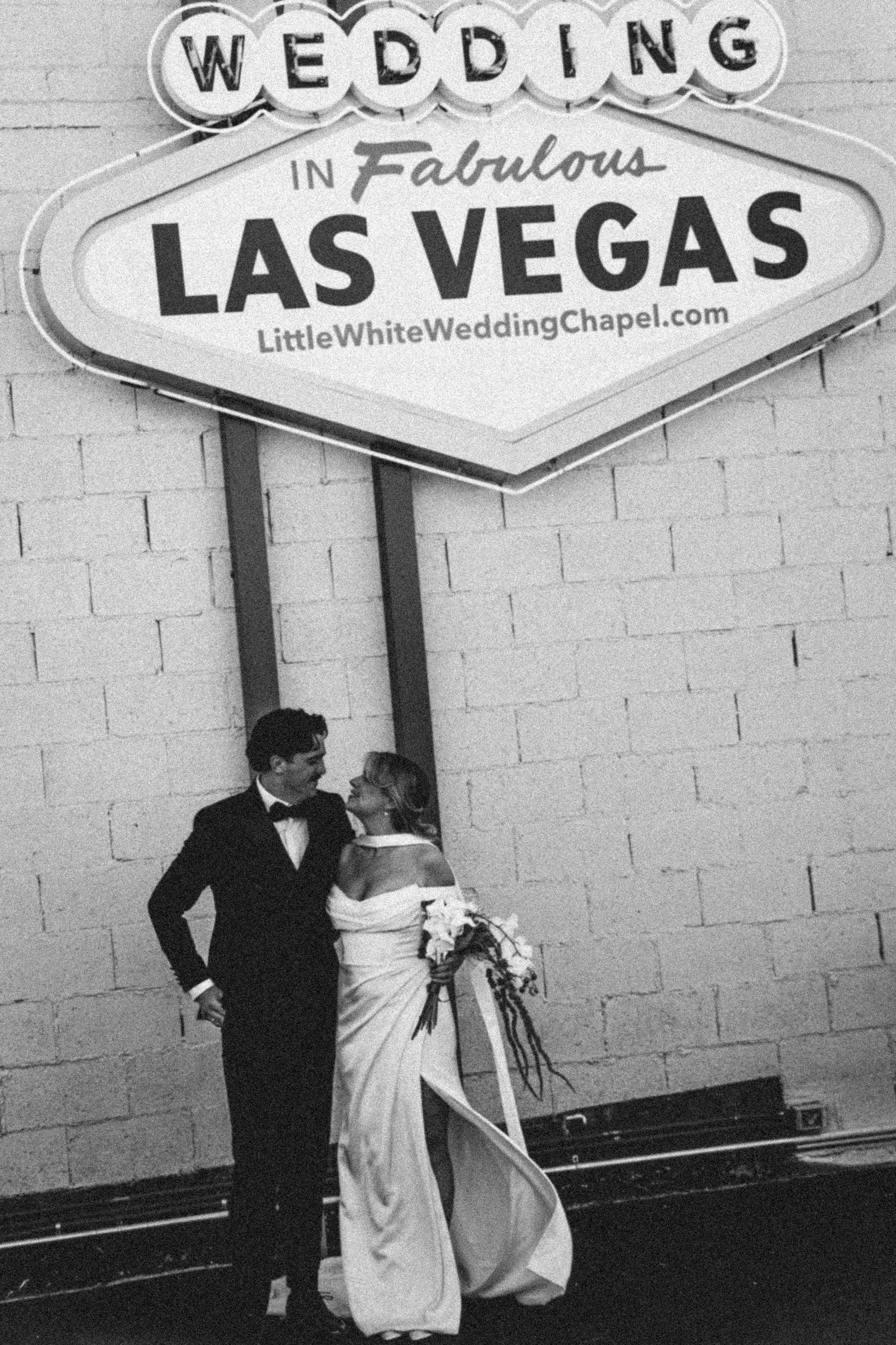 A newlywed couple standing together in front of a brick wall at a wedding reception, with a sign above that reads 'Wedding in Fabulous Las Vegas' and a website URL. The groom is in a tuxedo, and the bride is holding a bouquet of flowers and wearing a