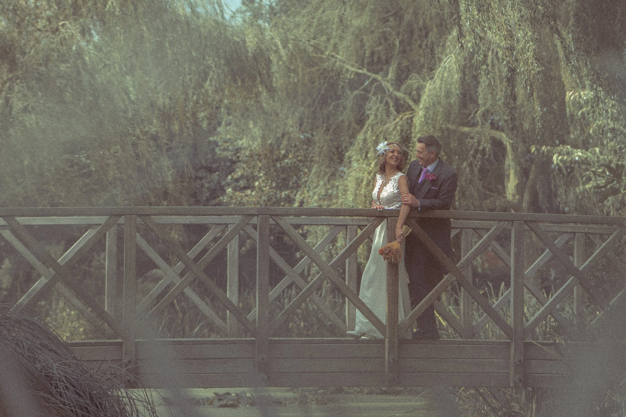 A newlywed couple stands on a wooden bridge in a lush forest, sharing a joyful moment together.
