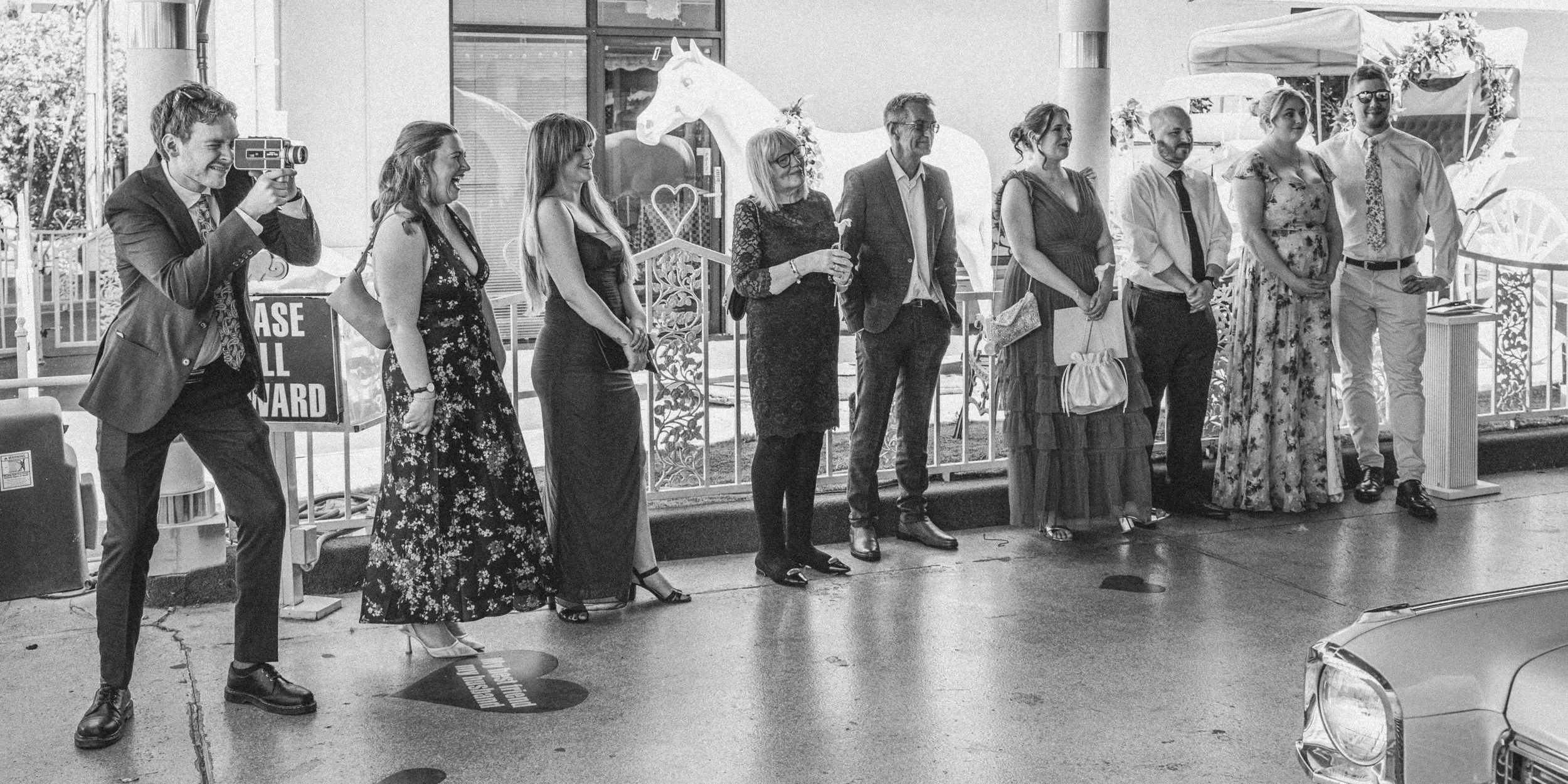 A group of nine people standing side by side outdoors, dressed in formal and casual attire, some smiling and others looking away. One person is taking a photo with a vintage camera. They are standing behind heart-shaped markers on the ground, and the