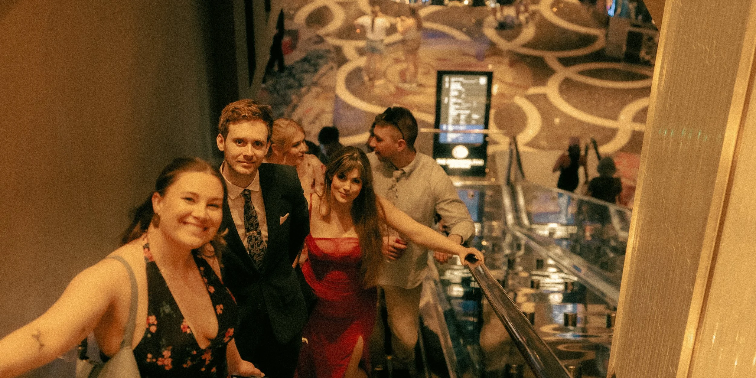 Group of people riding an escalator at a casino or hotel, with a patterned carpet in the background and a digital display board visible.