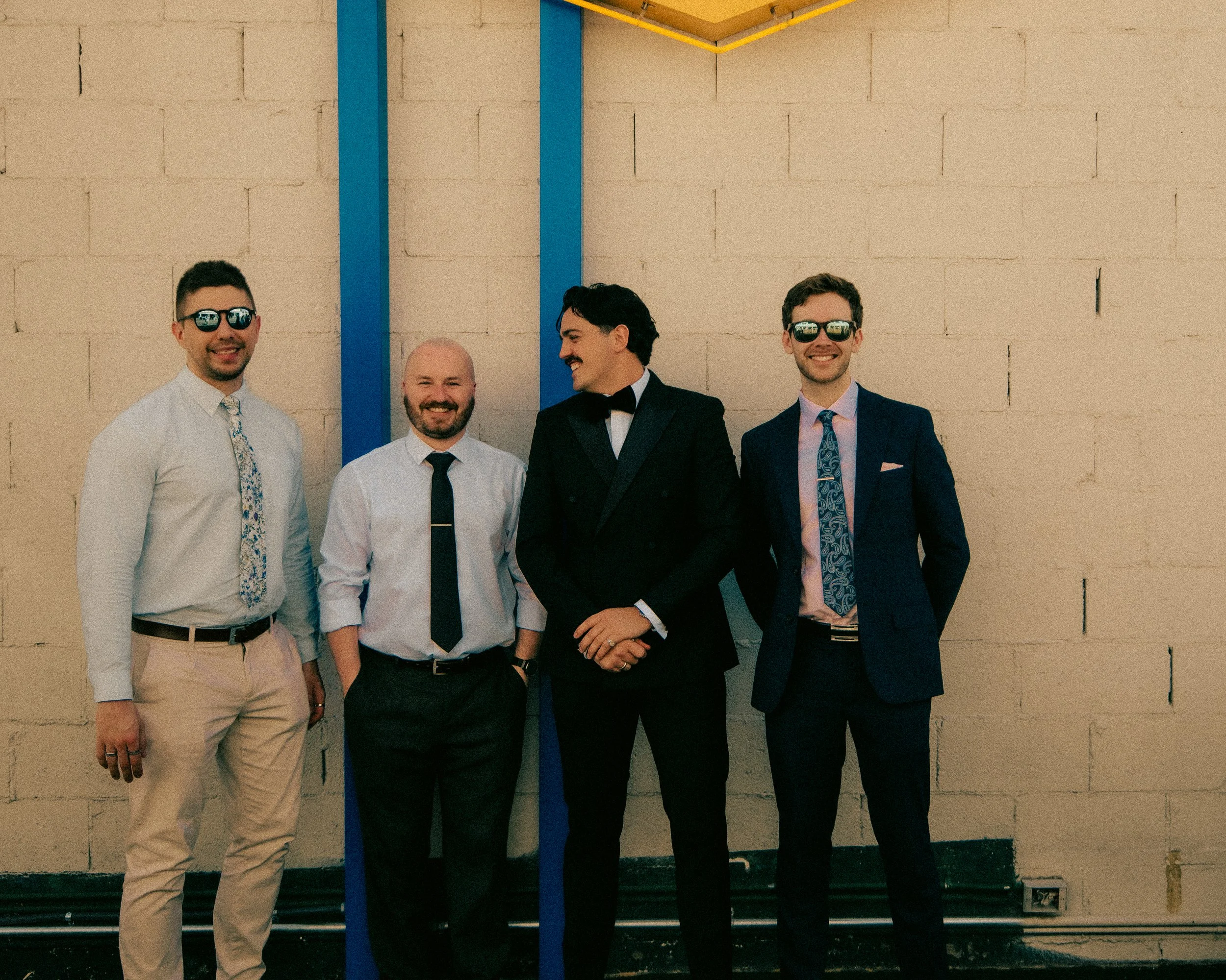 Four men in formal attire standing in front of a beige brick wall with a vertical blue line and yellow border at the top. Two of the men are wearing sunglasses, and all are smiling or smirking.