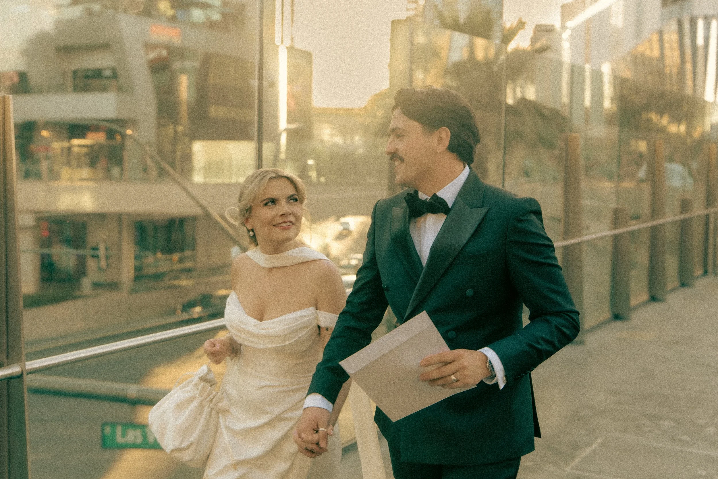 A bride and groom holding hands on a city street, walking past a glass building during sunset. The bride wears a white wedding dress with an off-shoulder style, and the groom wears a black tuxedo with a bow tie. They are smiling at each other.