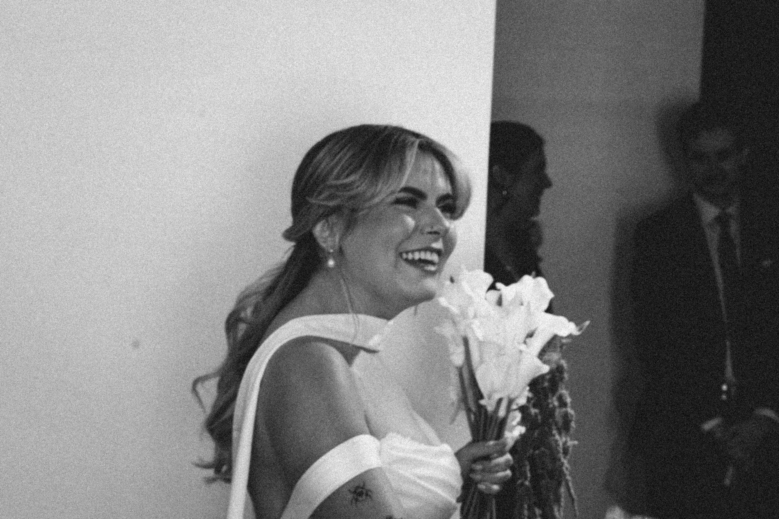 Black and white photo of a woman in formal attire smiling, holding a bouquet of flowers, standing next to a man in a tuxedo.