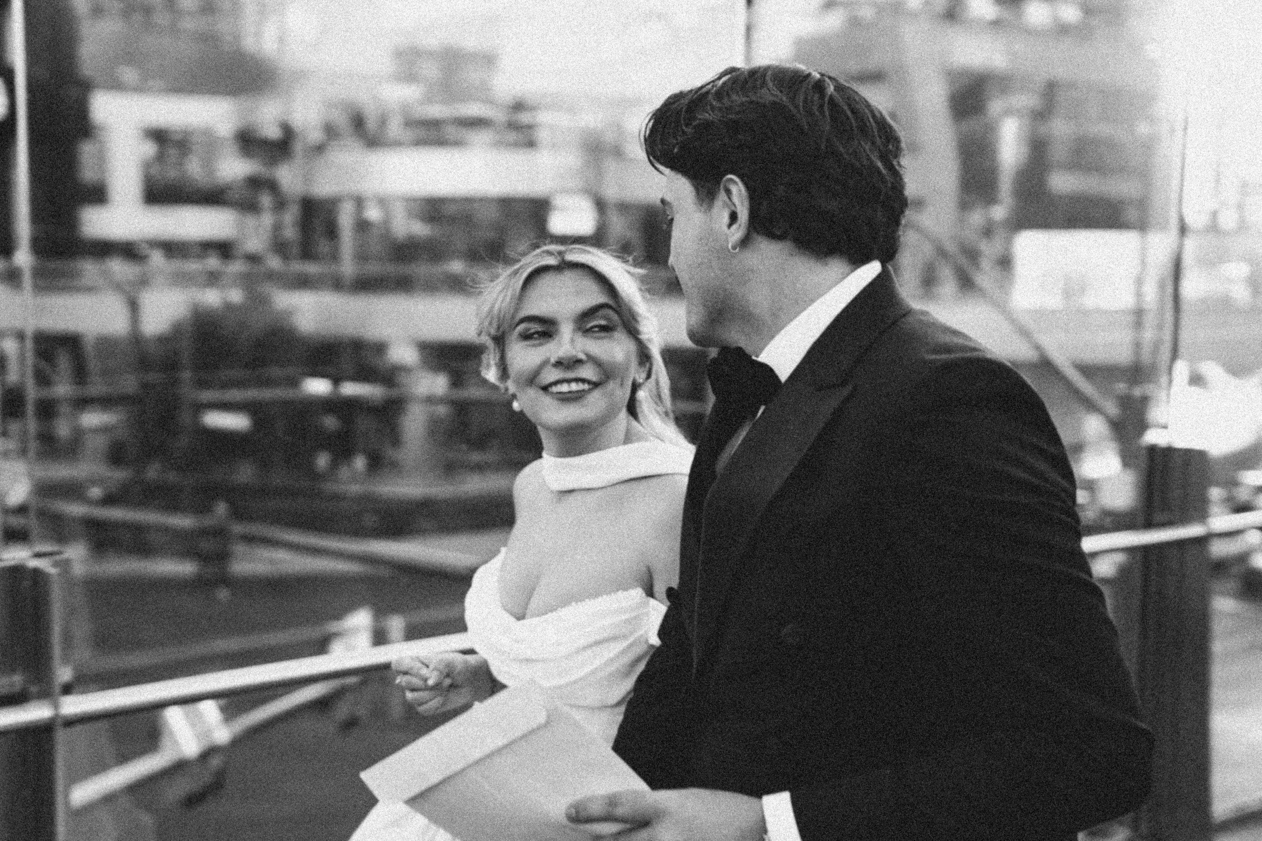 A black and white photo of a couple dancing together in a formal setting. The woman is smiling and wearing a strapless dress, while the man is dressed in a tuxedo. They are holding hands and gazing at each other, with an urban cityscape in the backgr