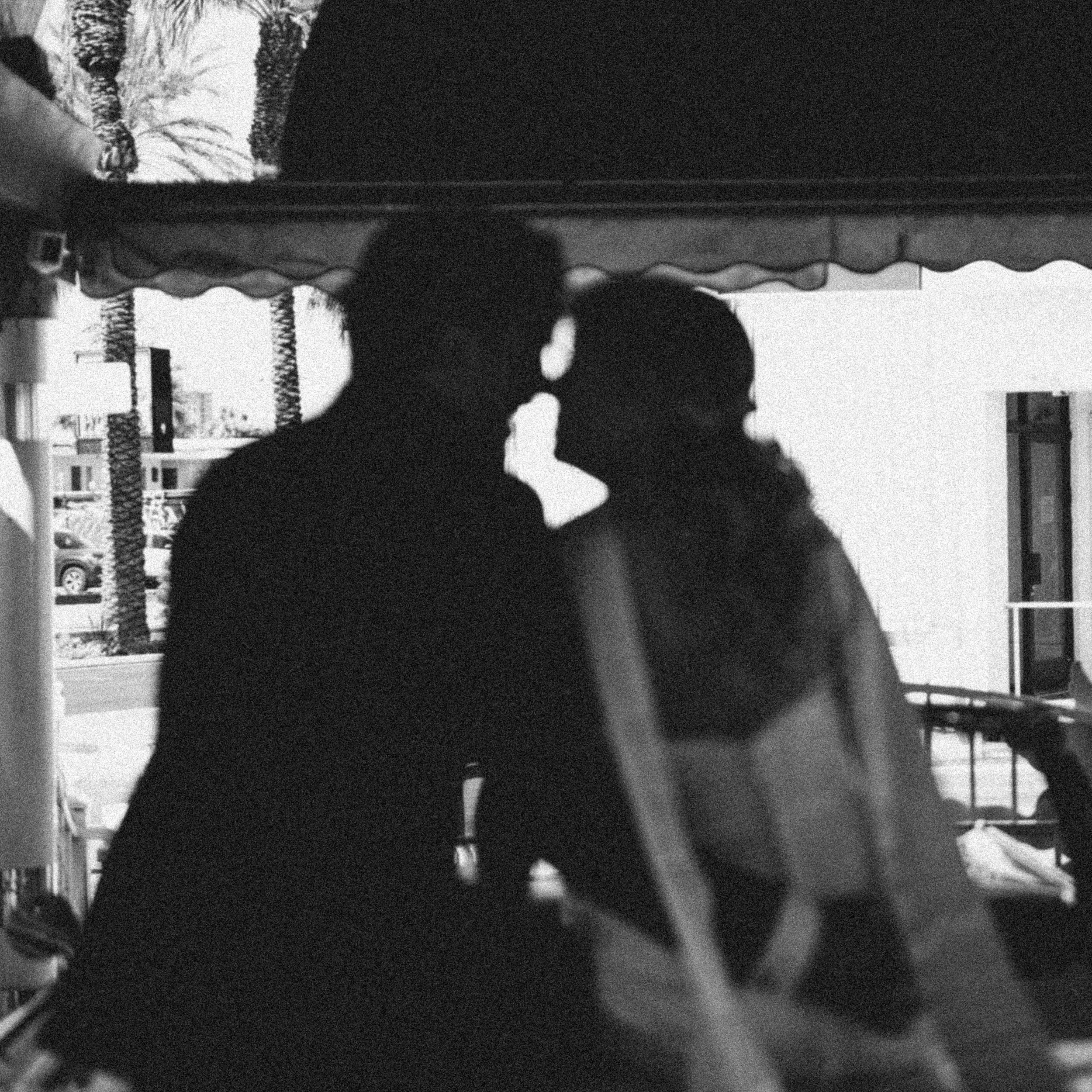 Silhouette of a couple kissing in front of a beachfront scene with palm trees and buildings in the background.