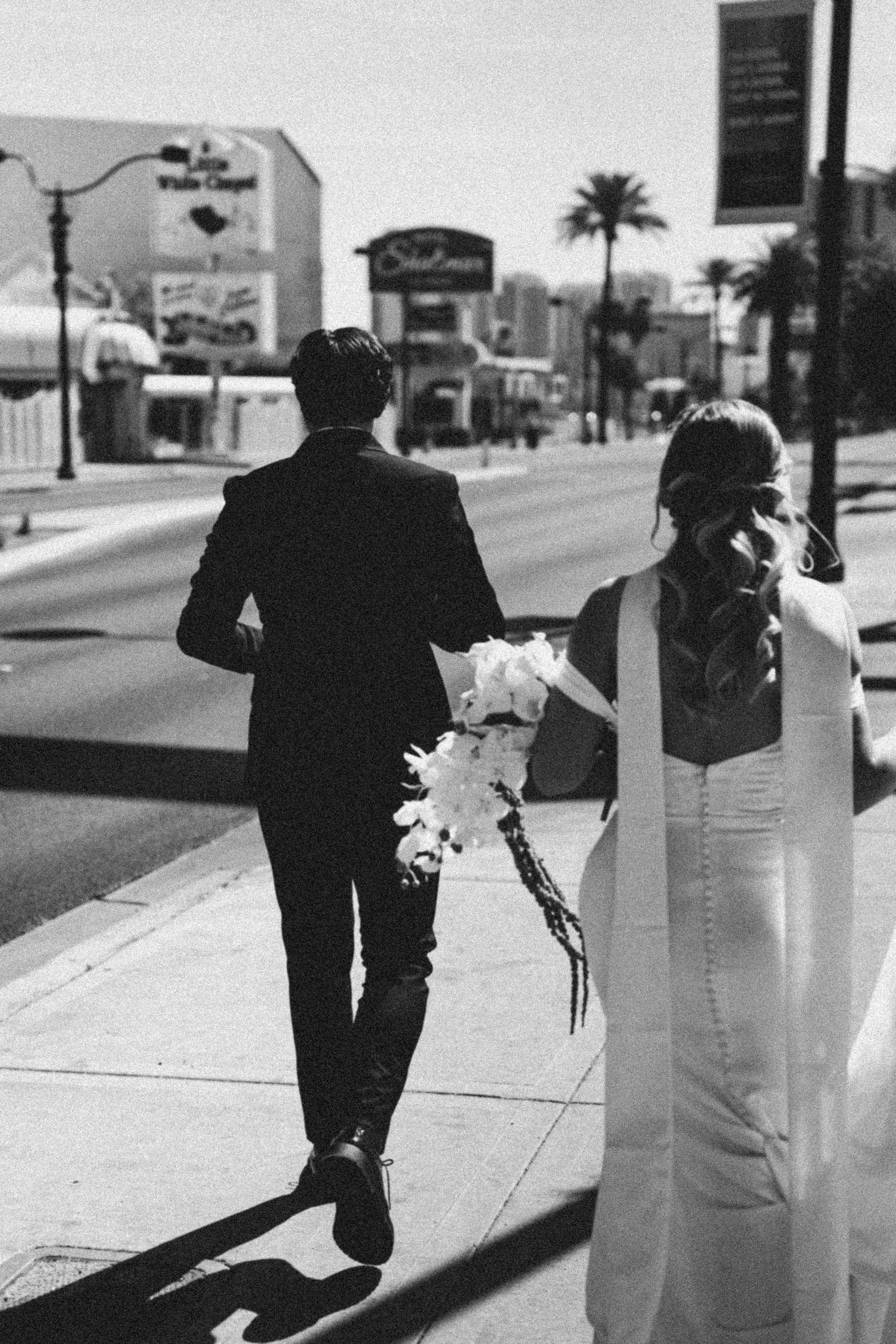 Black and white photo of a man and woman walking down a sidewalk on a sunny day. The man is dressed in a suit, holding flowers, and the woman has long hair tied back and is wearing a striped top. In the background, there are palm trees and buildings 