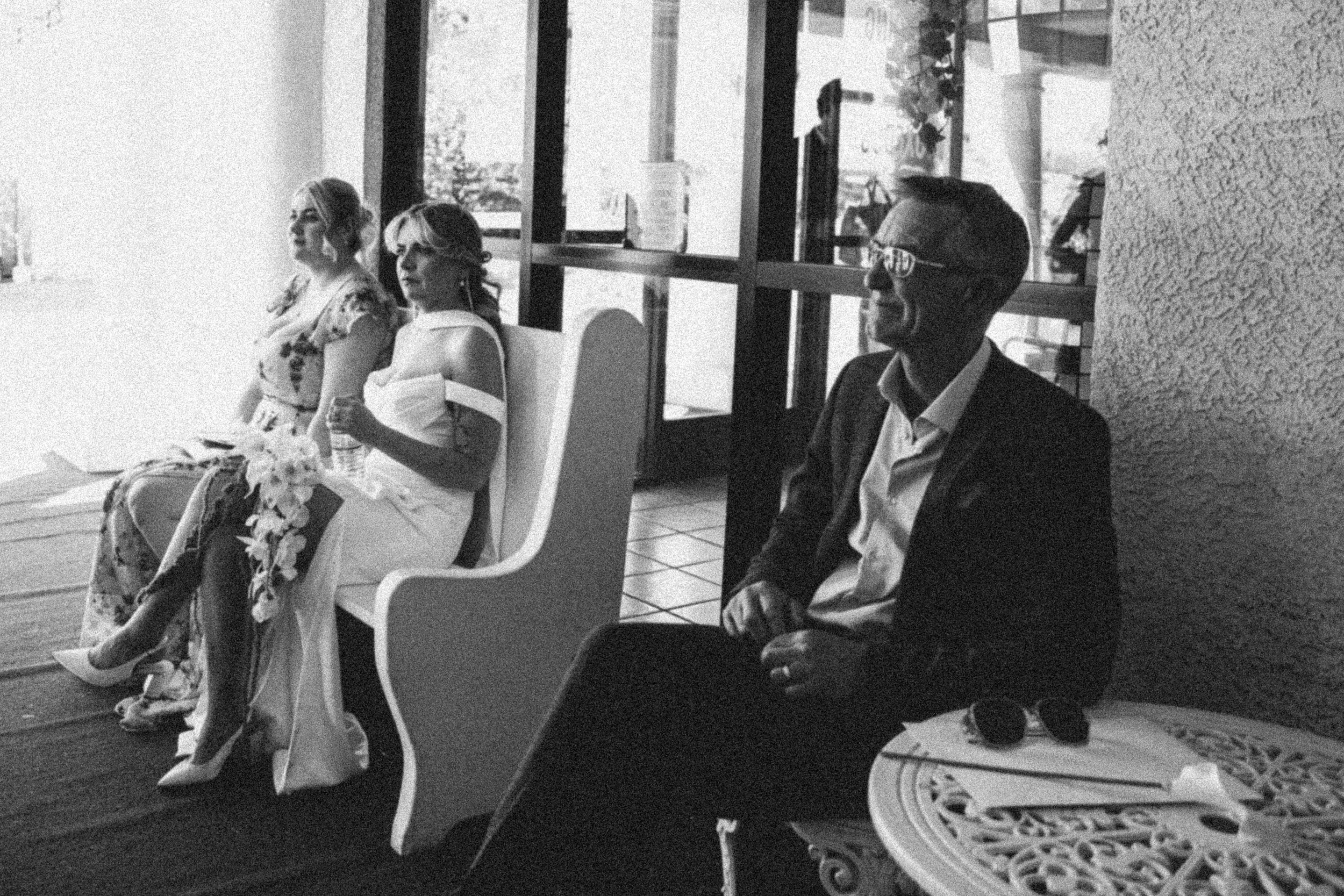 People sitting in a row at a formal indoor event, with a woman in a floral dress, a woman with glasses holding a piece of paper, and a man in a suit and glasses smiling, in front of large windows letting in natural light.
