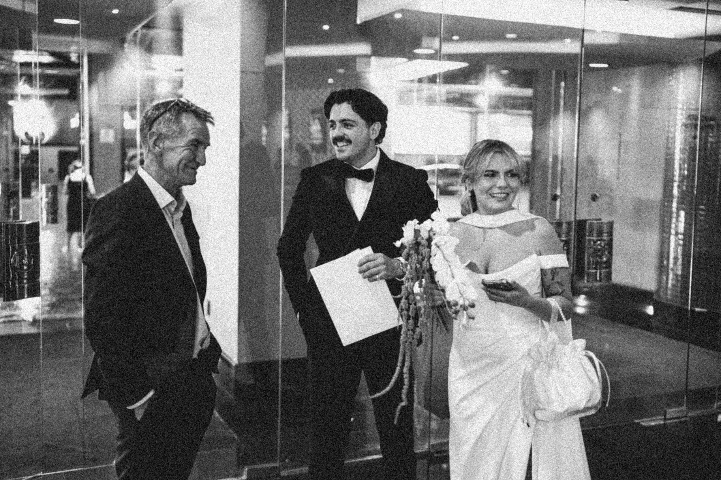 A black and white photo of a wedding scene inside a building, with three people: a bride in a wedding dress holding a bouquet, a man in a tuxedo, and another man in a suit, all smiling and engaging in conversation.