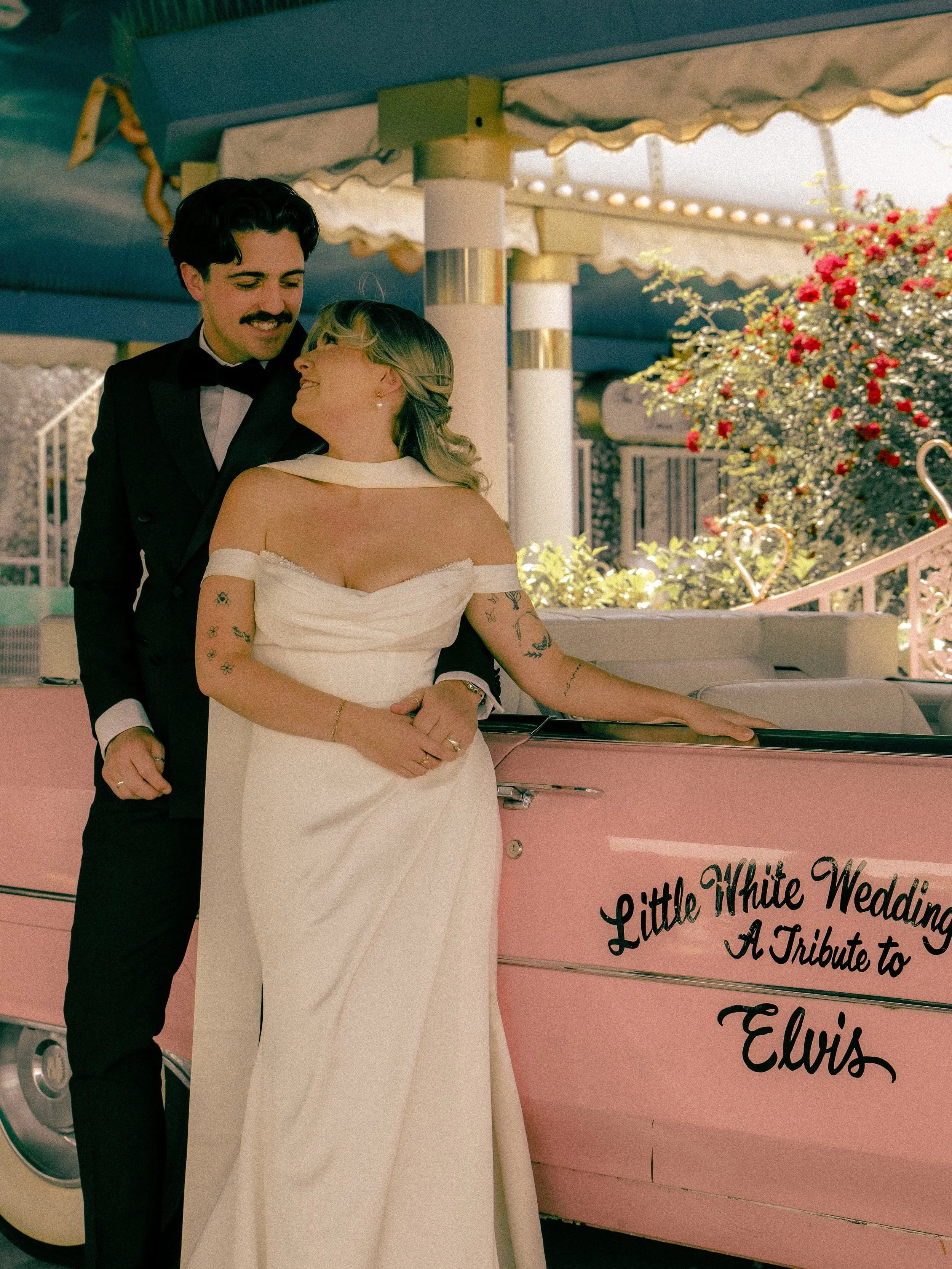 A couple dressed in wedding attire stands near a pink vintage teardrop trailer with black writing and flowers, under a striped awning with hanging lights and a bush with pink flowers in the background.
