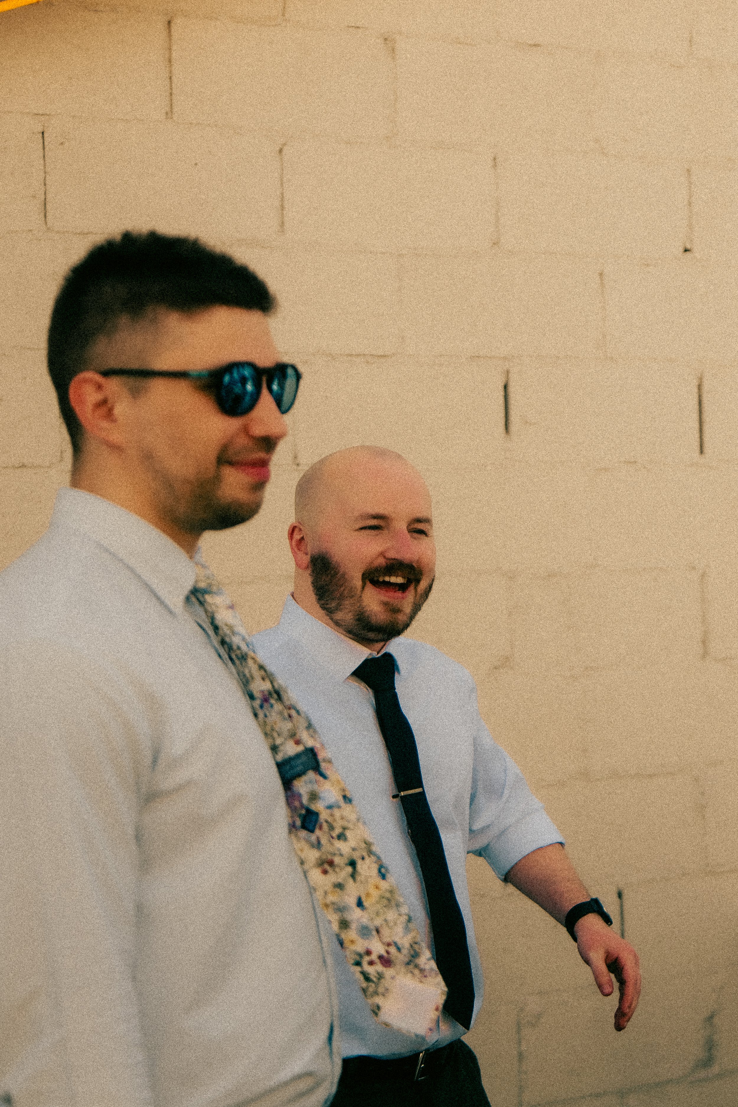 Two men dressed in formal shirts and ties standing in front of a beige brick wall, smiling and laughing.