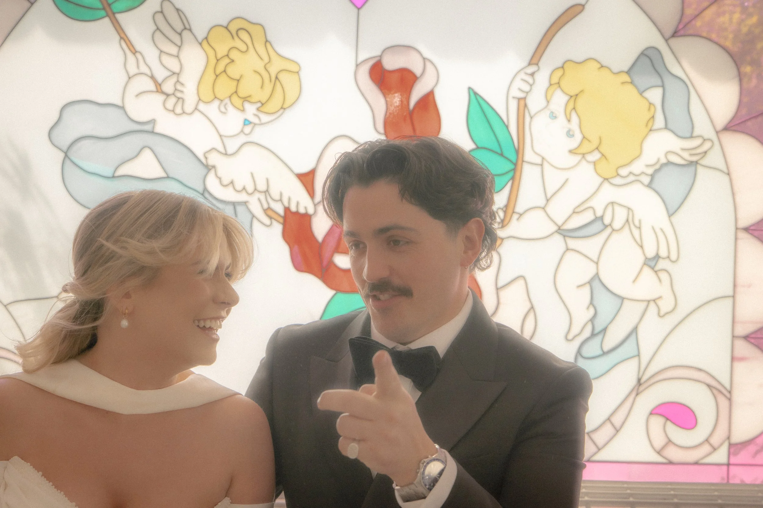 A bride and groom taking a selfie. The bride is smiling, and the groom is holding a phone. They are dressed formally. There is a decorative background with cartoon cherubs and floral designs.