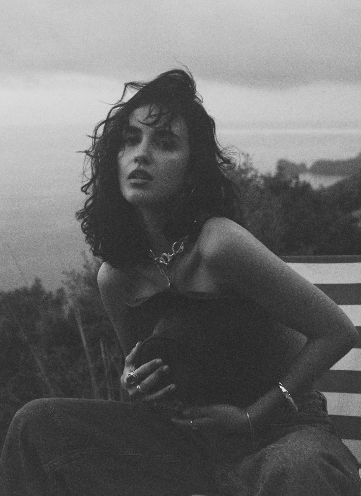 A black-and-white photo of a woman with curly hair, wearing a dark off-shoulder top and jewelry, sitting outdoors against a backdrop of cloudy sky and trees.