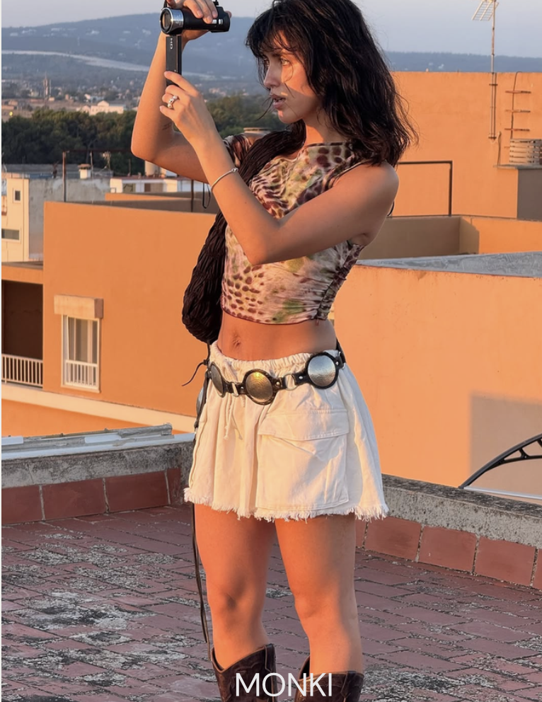 Young woman with black hair taking a photo with a small camera on a rooftop at sunset. She wears a colorful tie-dye crop top, white frayed shorts, a belt with large reflective circular buckles, and dark boots. Cityscape with buildings and hills in the background.