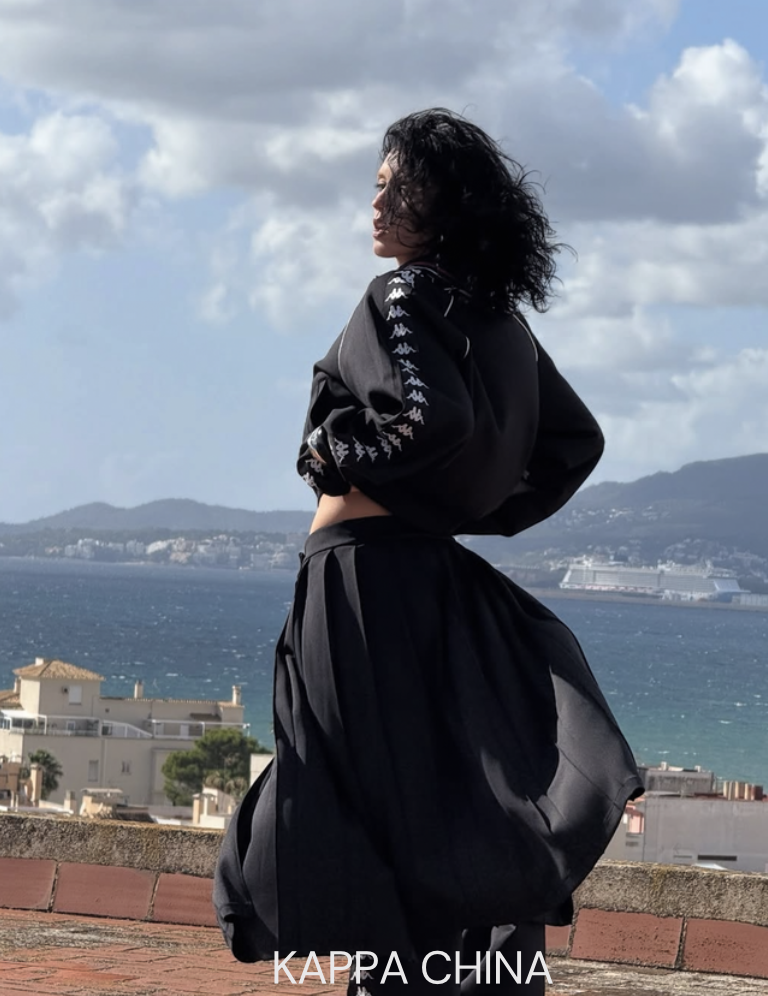 Woman with curly black hair wearing a black jacket and skirt standing on a brick wall by the sea with mountains and cloudy sky in the background.