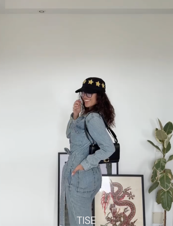 A woman wearing a denim jumpsuit, black cap with yellow stars, and glasses, smiling and posing indoors near framed artwork and a tall green plant.
