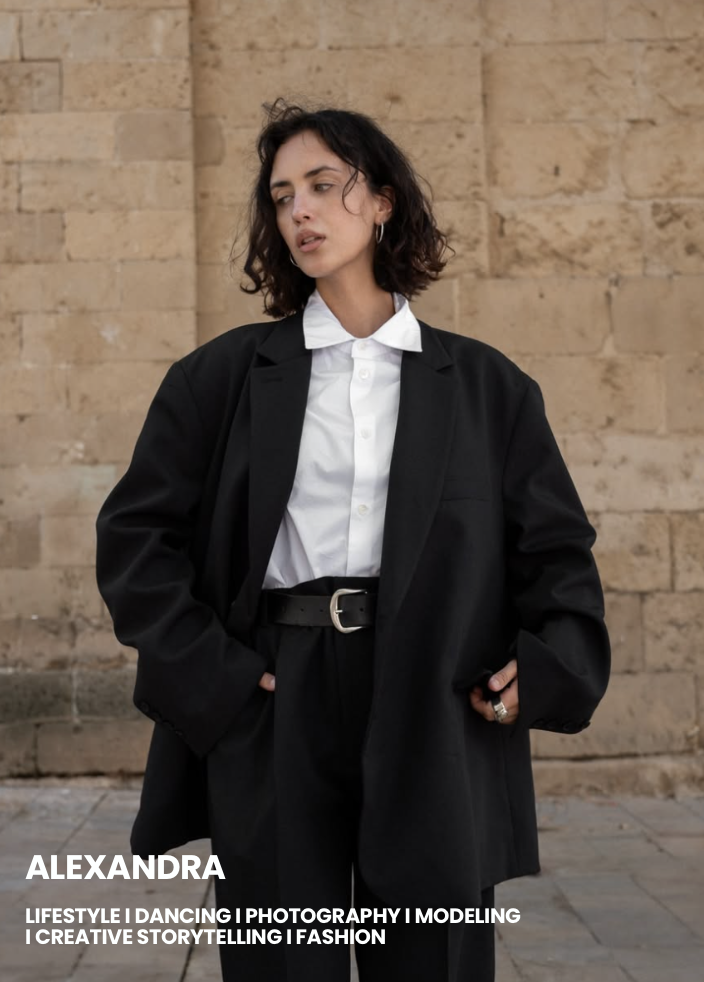 A woman with short curly dark hair wearing a white button-up shirt, black blazer, and black pants with a belt, standing against a beige stone wall.