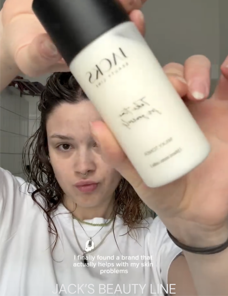 Young woman in a white shirt holding a bottle of skincare product towards the camera in a bathroom.