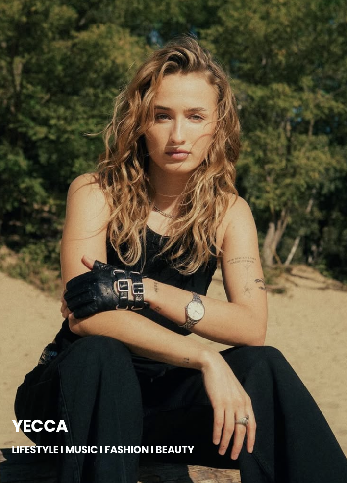 A young woman with wavy blonde hair and light skin sitting outdoors in a natural setting with green trees in the background. She is wearing a sleeveless black top, black pants, a watch, and a glove on one hand, with her arms crossed and a serious expression.