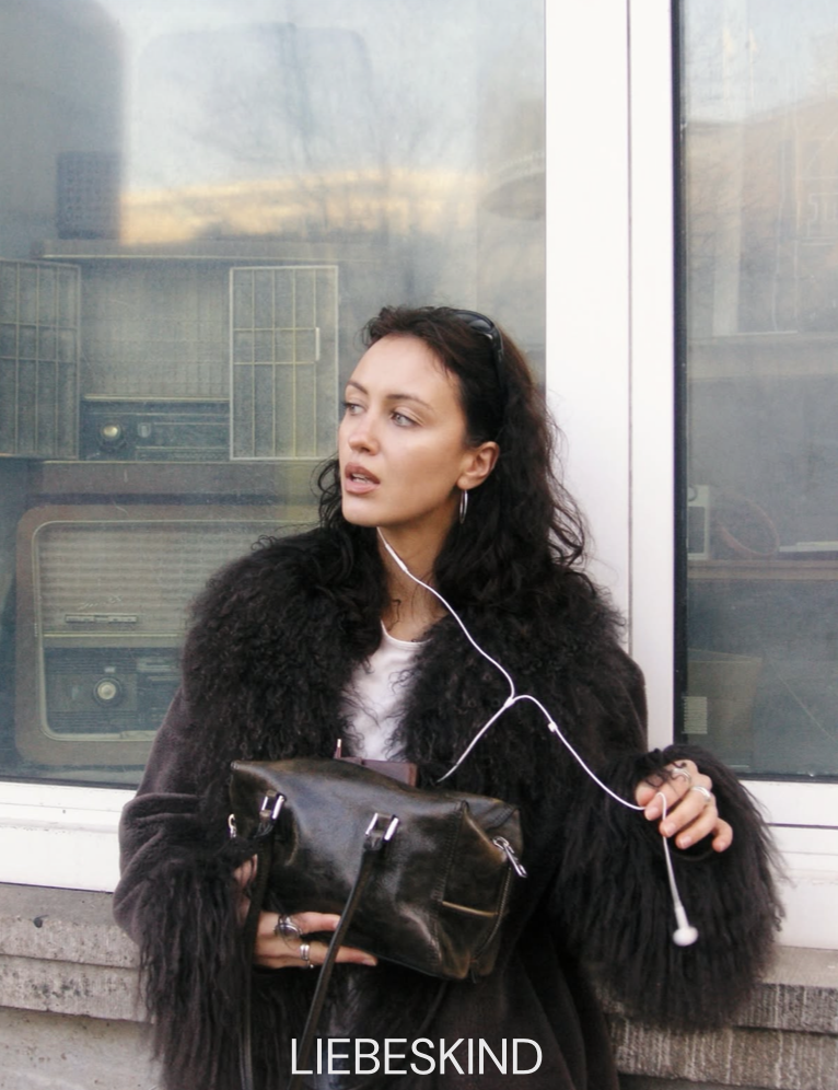 A woman with dark curly hair, wearing a fuzzy black coat, holding a black handbag, standing in front of a reflective glass window, with earphones in her ears.
