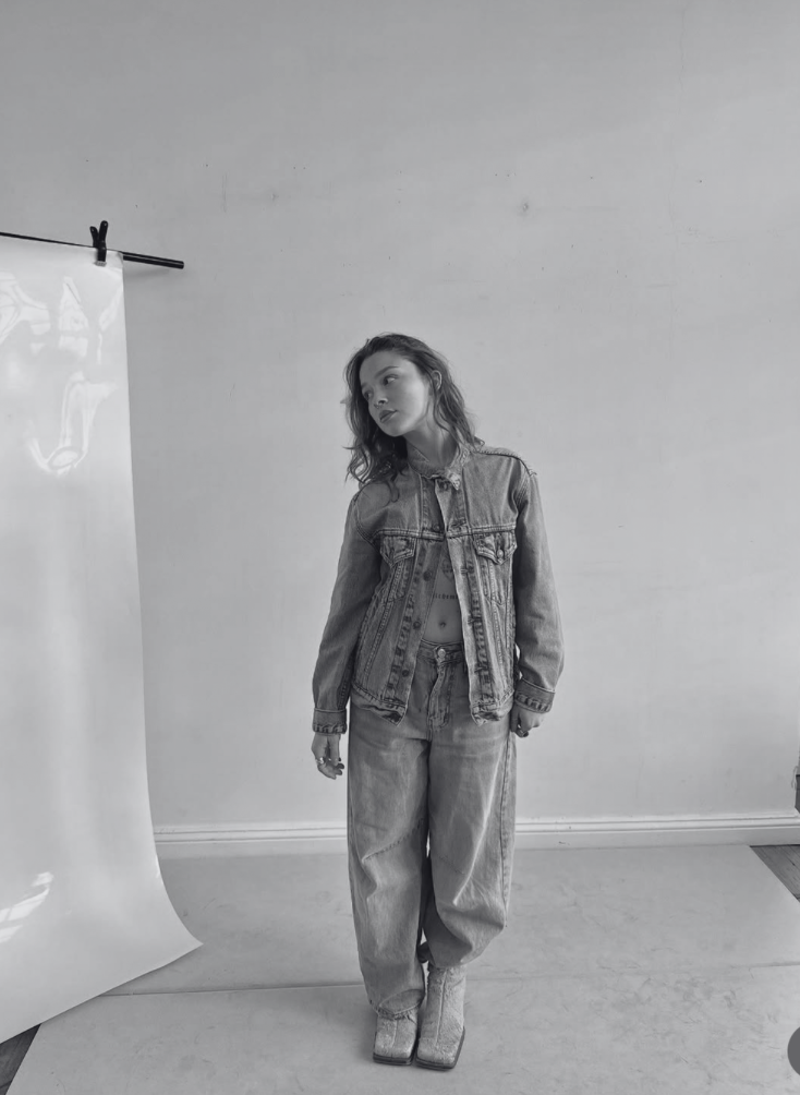 A young woman with wavy hair standing indoors against a plain wall, dressed in a denim jacket, high-waisted jeans, and cowboy boots.