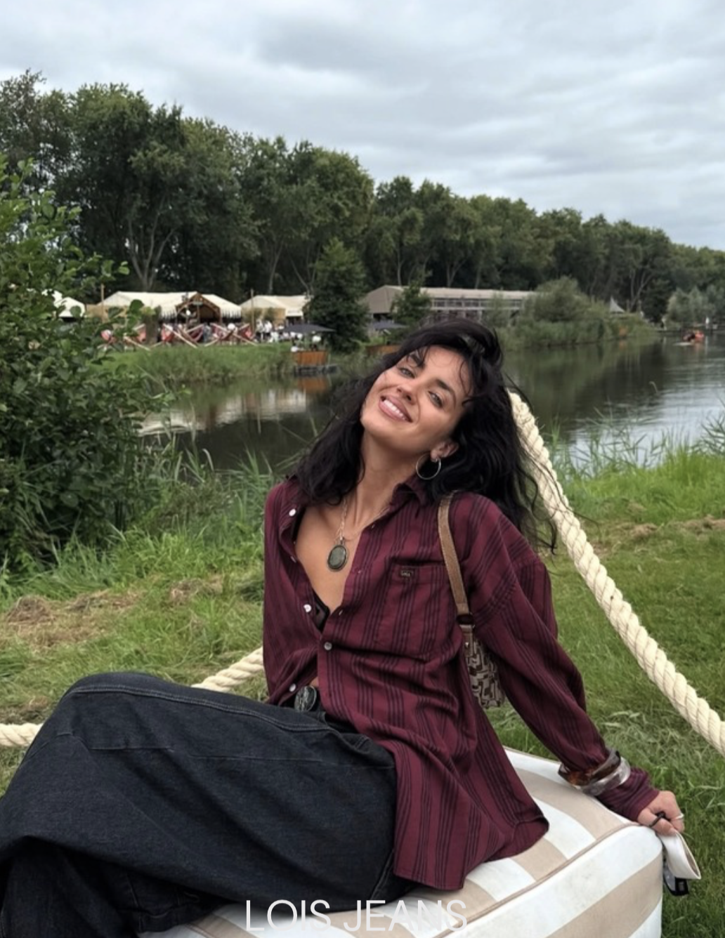 A woman with dark hair and a smile, wearing a red plaid shirt and black jeans, sitting on a swing by a river with trees and tents in the background.