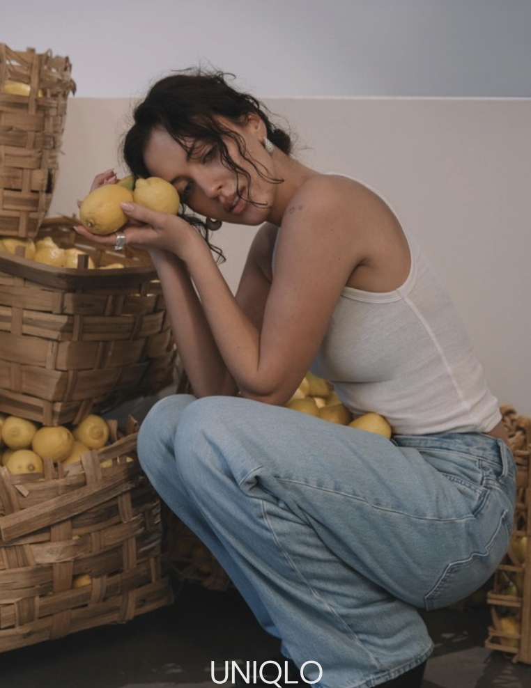 A woman with curly dark hair in a white tank top and light blue jeans sitting next to baskets of lemons, holding two lemons close to her face.